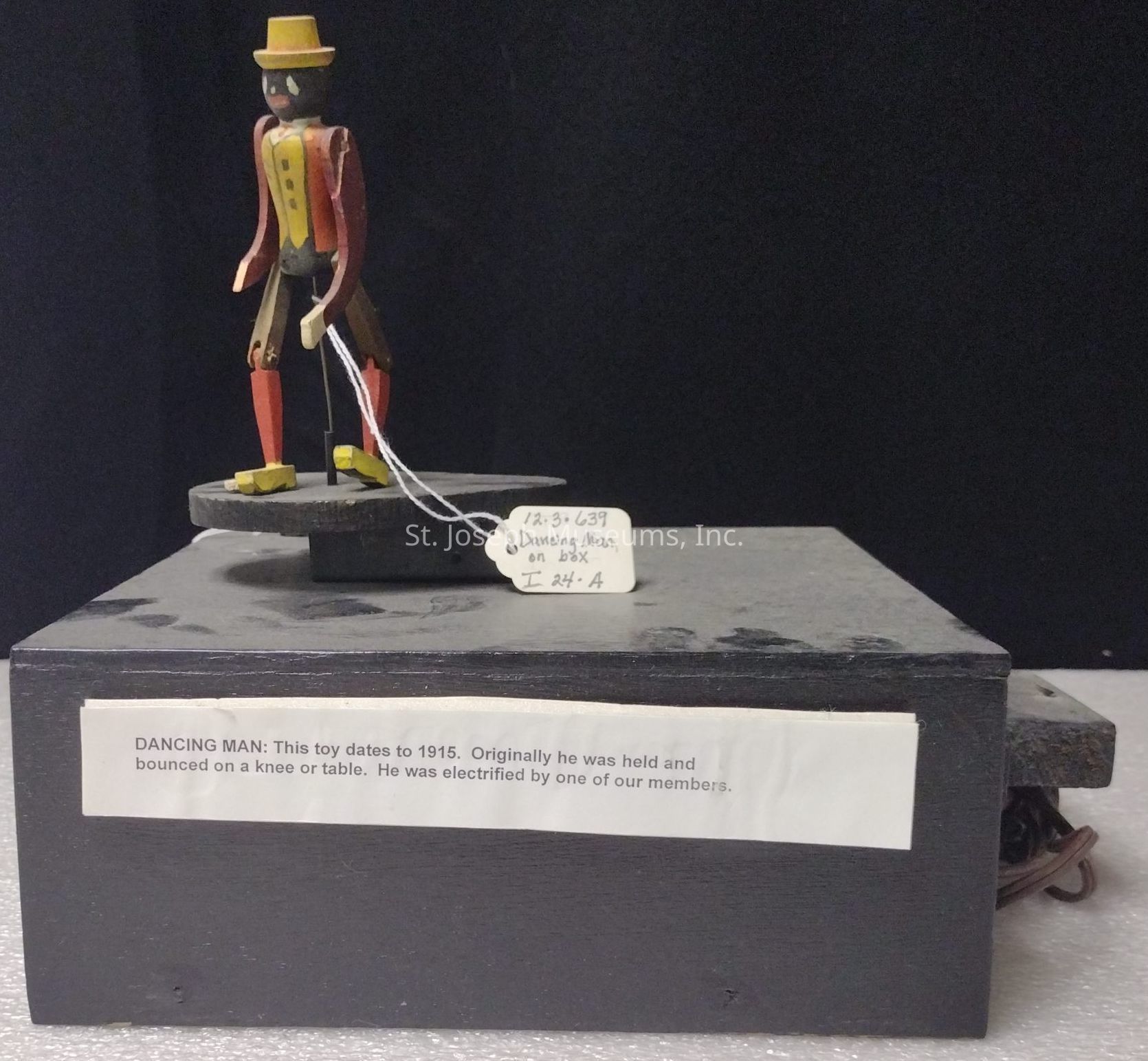 The image depicts a vintage toy labeled "Dancing Man," which dates back to 1915. The toy consists of a small wooden figure, painted in bright colors including yellow, red, and black, adorned with a hat and positioned on top of a black box. The figure appears to be mechanized, as suggested by a description that states "Originally he was held and bounced on a knee or table. He was electrified by one of our members." The tag with handwritten text attached to the toy reads "Dancing Man on Box," accompanied by numbers and possibly a catalog reference. This piece seems to be showcased for its historical value, offering a glimpse into early 20th-century toy-making and entertainment.