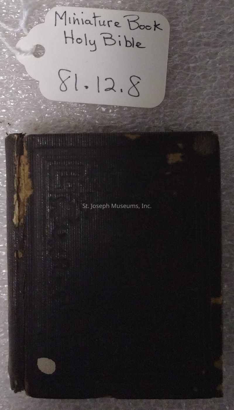 The image depicts a small, worn-out book with a dark cover, identified by a handwritten tag lying beside it that reads "Miniature Book Holy Bible" along with the number "81.12.8." The cover appears to have some peeling and faded spots, suggesting age or frequent use. The book is placed on a textured surface, possibly bubble wrap, indicating it might be part of a collection or an item being stored carefully. The compact size of the book and the presence of a descriptive tag suggest its significance or uniqueness, possibly as a collectible or an antique item.