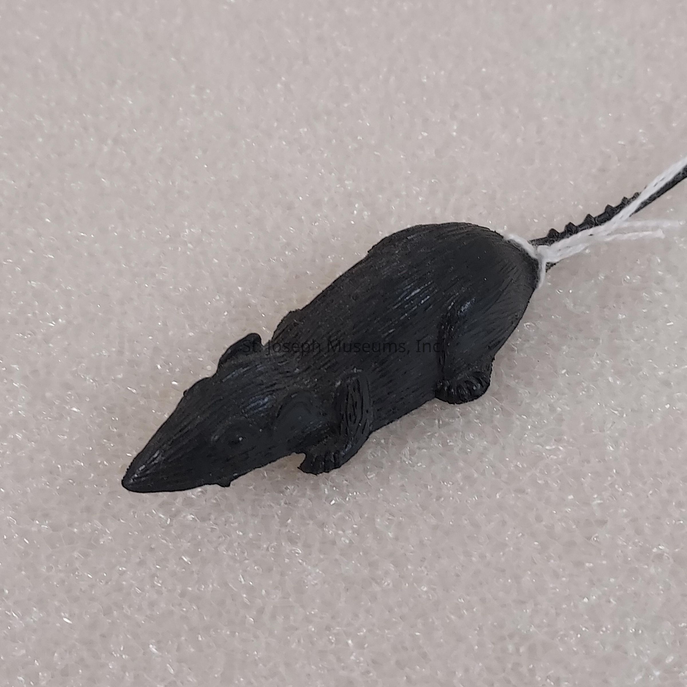 The image depicts a small black figurine of a rat placed on a textured, light-colored surface. The figurine has a detailed design with distinct features such as a long pointed snout, rounded ears, and a long thin tail. The tail is adorned with a white string that is attached near its base, hinting at the possibility that this object might be used as an accessory or decoration, perhaps a keychain or a small ornament. The smooth and slightly reflective surface of the rat gives it a plastic-like appearance, suggesting it might be made of a lightweight material such as molded plastic or resin. The background surface has a rough, porous texture, likely indicating a type of foam or fabric. The contrast between the black figurine and the light background highlights the rat, making it a focal point of the image, as the lighting enhances the shadows on the textured surface, emphasizing the rat's form and details.