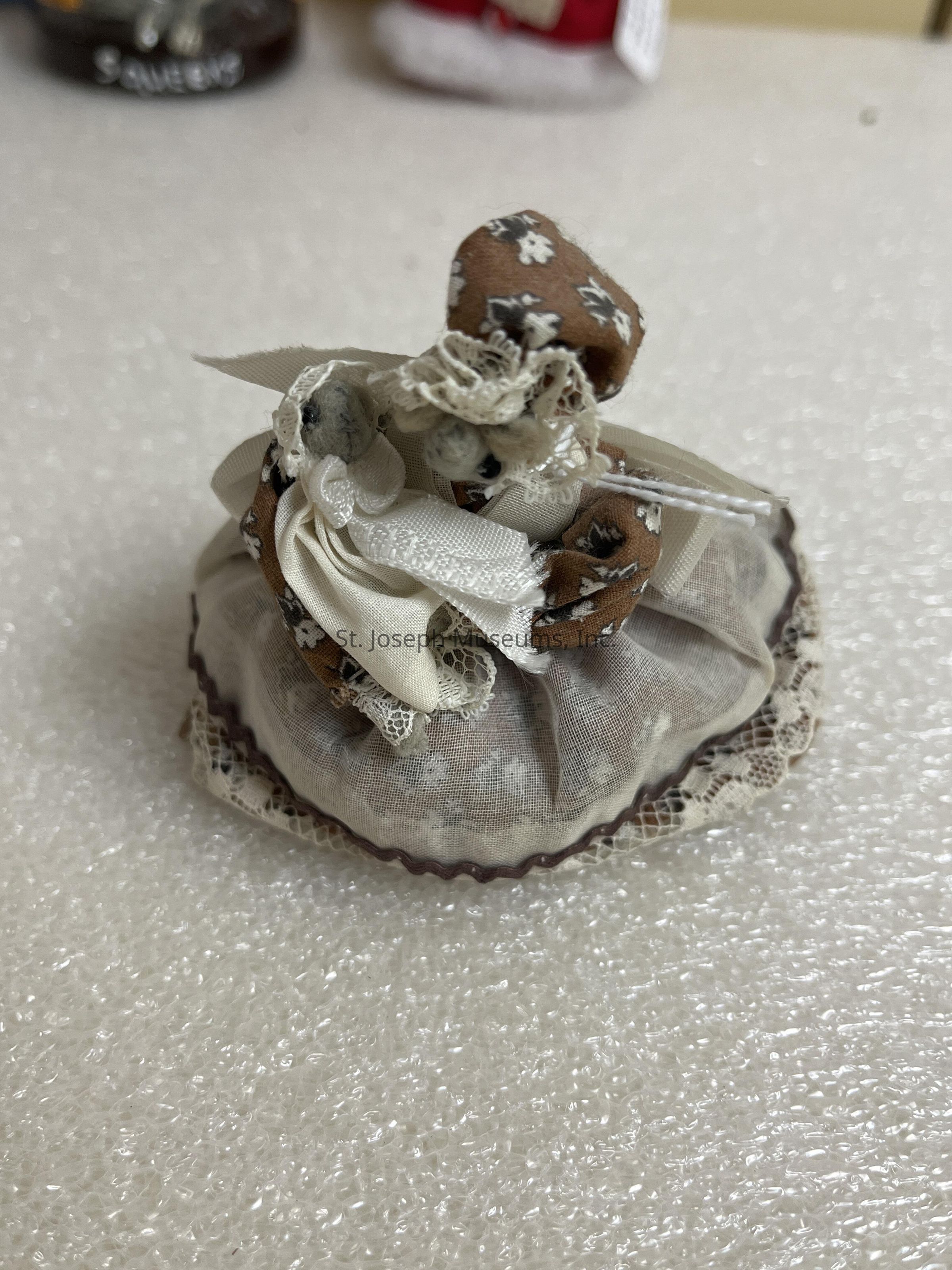 The image features a small, handcrafted doll made of fabric and lace. The doll is adorned with a brown and white patterned fabric, which covers its head and forms part of its attire. It is embellished with lace details and a white ribbon, adding a touch of elegance. The base of the doll appears to be a soft, round cushion, trimmed with lace, giving it a vintage appearance. The doll is placed on a textured, bubble-wrap-like surface, possibly for protection or display. The background is softly blurred, emphasizing the intricate details of the doll itself.