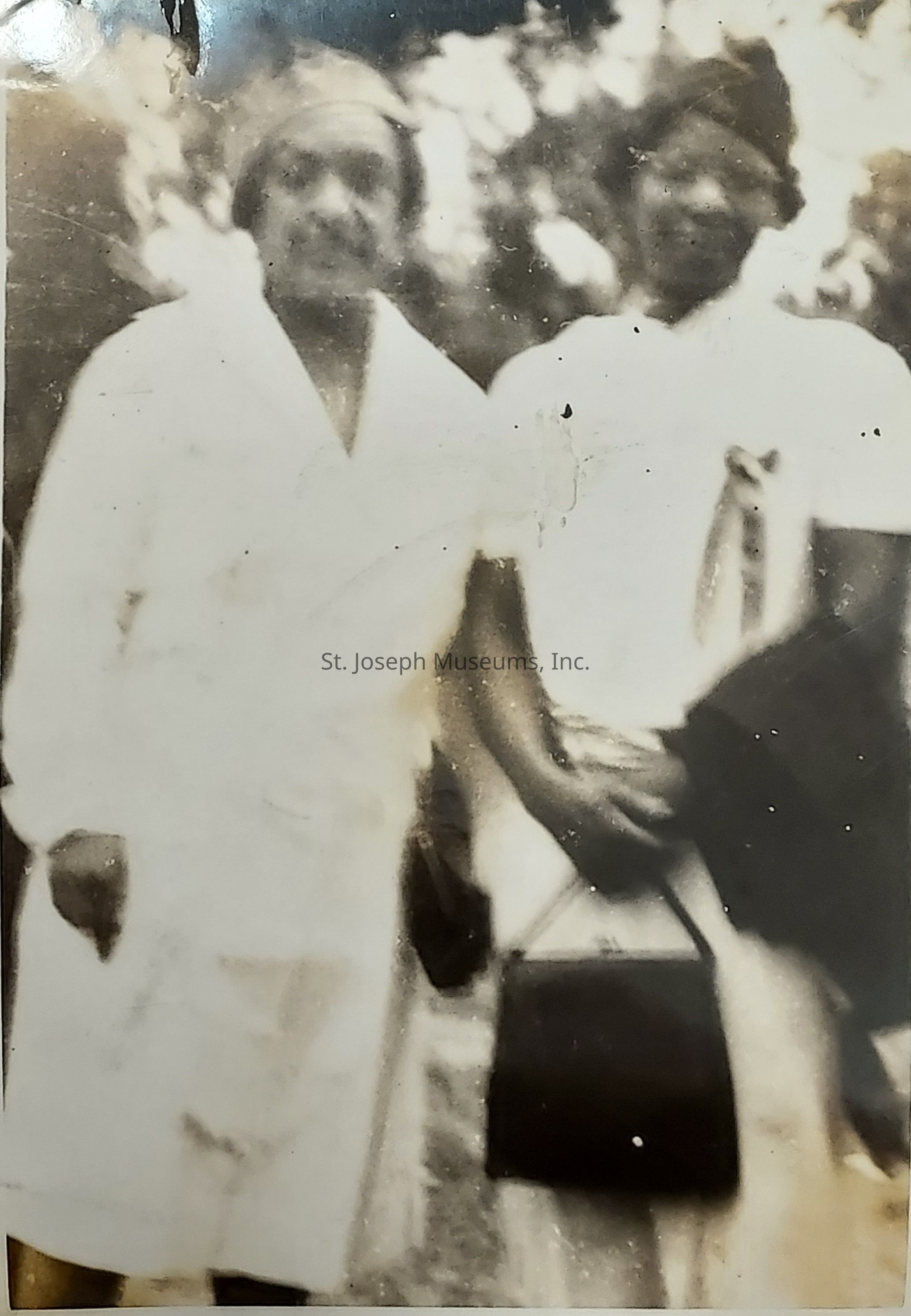The image depicts an old black and white photograph featuring two individuals standing closely together, with blurred and slightly faded details. Both are wearing light-colored clothing with the person on the left appearing to be in a dress or coat and possibly wearing a headscarf or hat. The person on the right is holding a small handbag and seems to be wearing a blouse with some kind of tied scarf or accessory. The background is indistinct but suggests an outdoor setting, likely with trees or foliage, given the shadows and shapes. The photograph has some visible wear and tear, with creases and marks, indicating its age and the possibility of it being a cherished keepsake or a snapshot from an earlier time. The expressions and specific features of the individuals are not clear, but they seem comfortable and familiar with each other, suggesting a relationship of friendship or family. Overall, this photograph gives a nostalgic glimpse into a moment captured in time, conveying warmth and a sense of history.