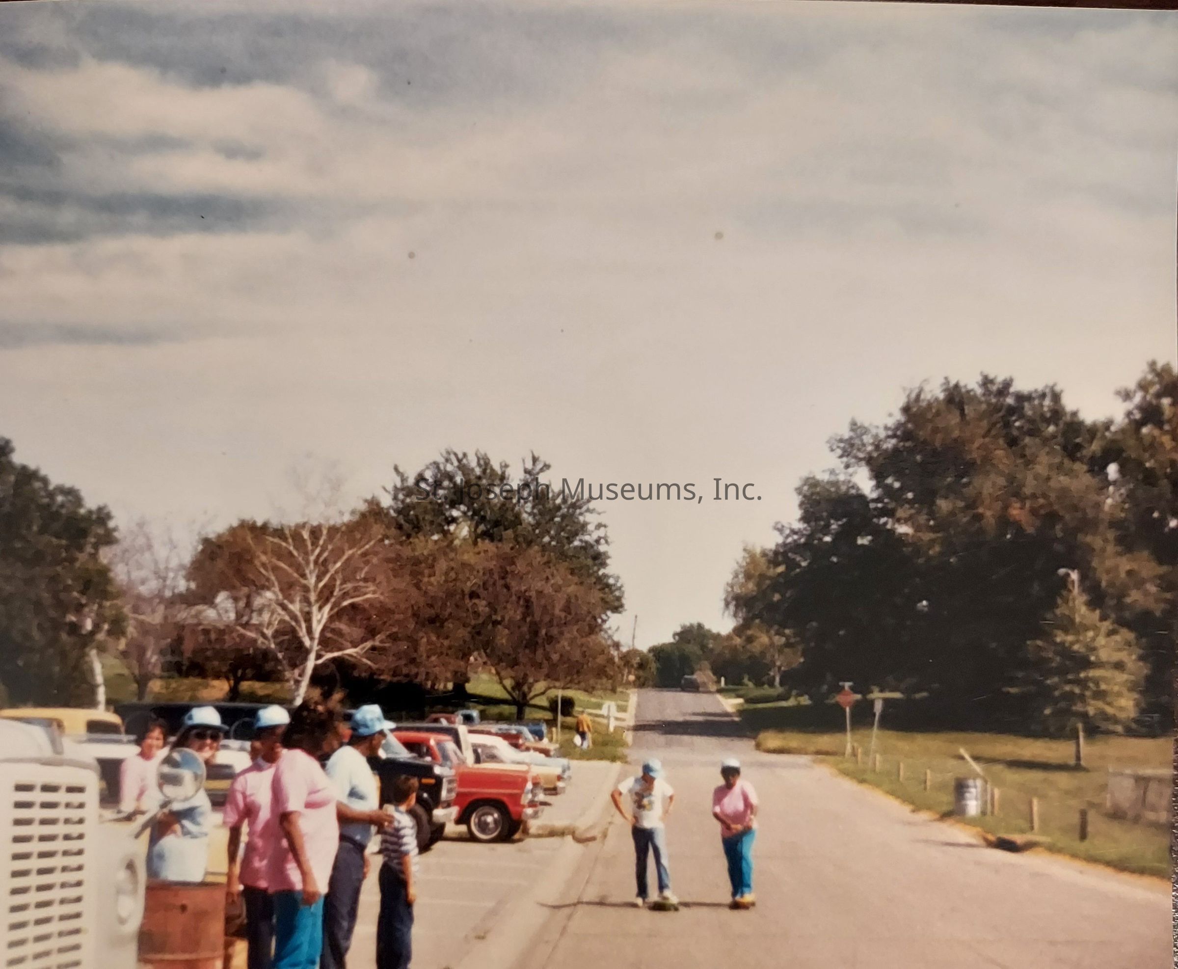 The image depicts a group of people gathered outdoors on a clear day, with some individuals standing near a line of parked vehicles on a wide street bordered by grass and large trees. On the left side of the street, a few people wearing light blue caps and pink shirts seem to be part of a group, possibly engaged in a communal activity or event. In the foreground, two young individuals with helmets are seen standing on skateboards in the middle of the road, suggesting a casual, leisurely atmosphere. The trees lining the street are lush and green, indicating a serene neighborhood setting. In the background, the street appears to extend into the distance, flanked by fences and more trees, giving the scene a peaceful suburban feel. A mix of vehicle colors, including red and yellow, stand out against the muted tones of the other elements, adding a splash of vibrancy to the scene. The sky is partly cloudy, suggesting favorable weather conditions for outdoor activities.