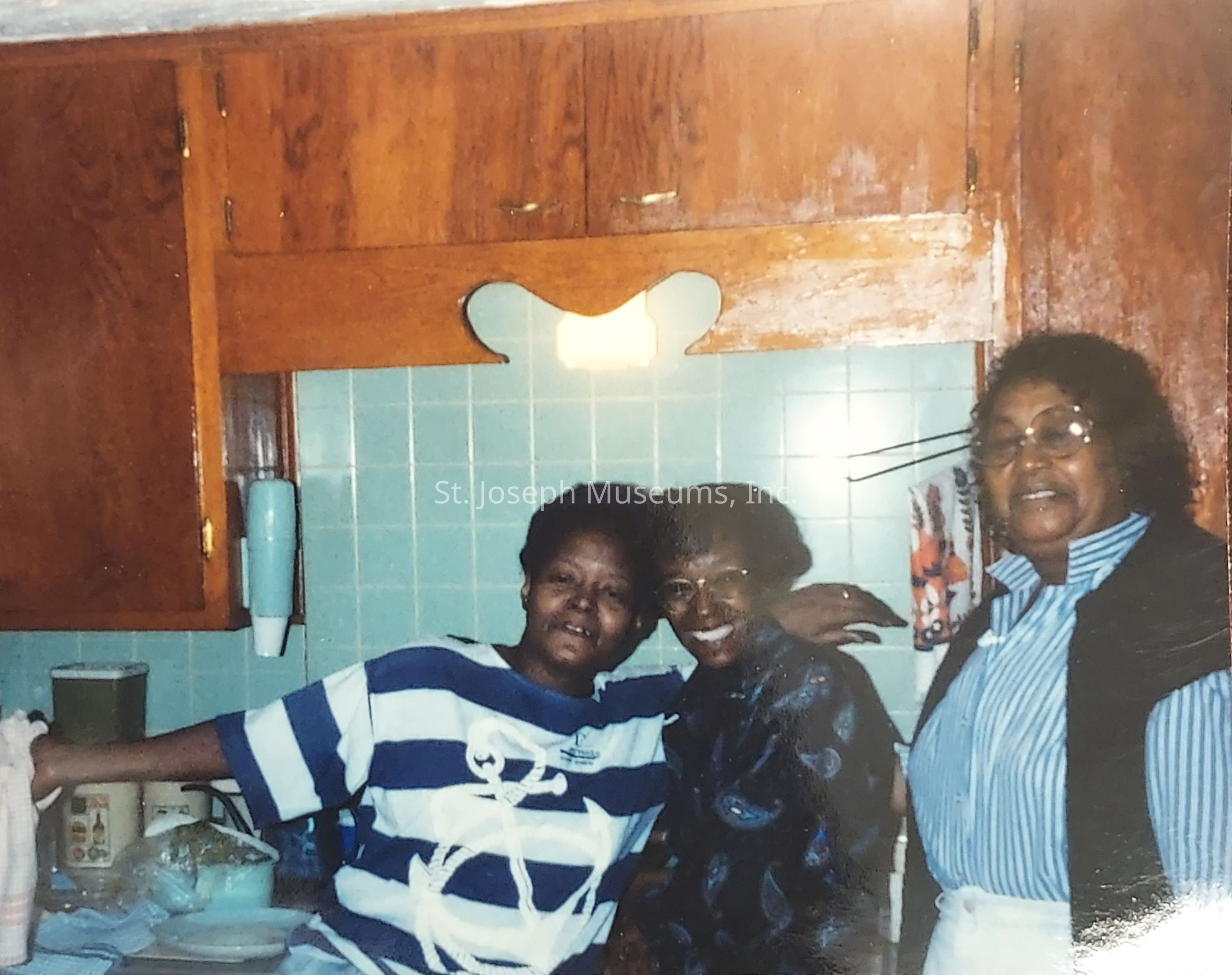 In a warm kitchen setting with light blue tiled wall decoration and wooden cabinets, three women stand closely together expressing a sense of camaraderie and happiness; the woman on the left leans casually against the countertop wearing a navy and white striped shirt, the middle woman, in a patterned shirt and glasses, smiles broadly with her arm around the others, and the woman on the right, wearing a blue-striped shirt with a vest, also smiles warmly, suggesting a moment of shared joy and possibly capturing a memorable family gathering or celebration.