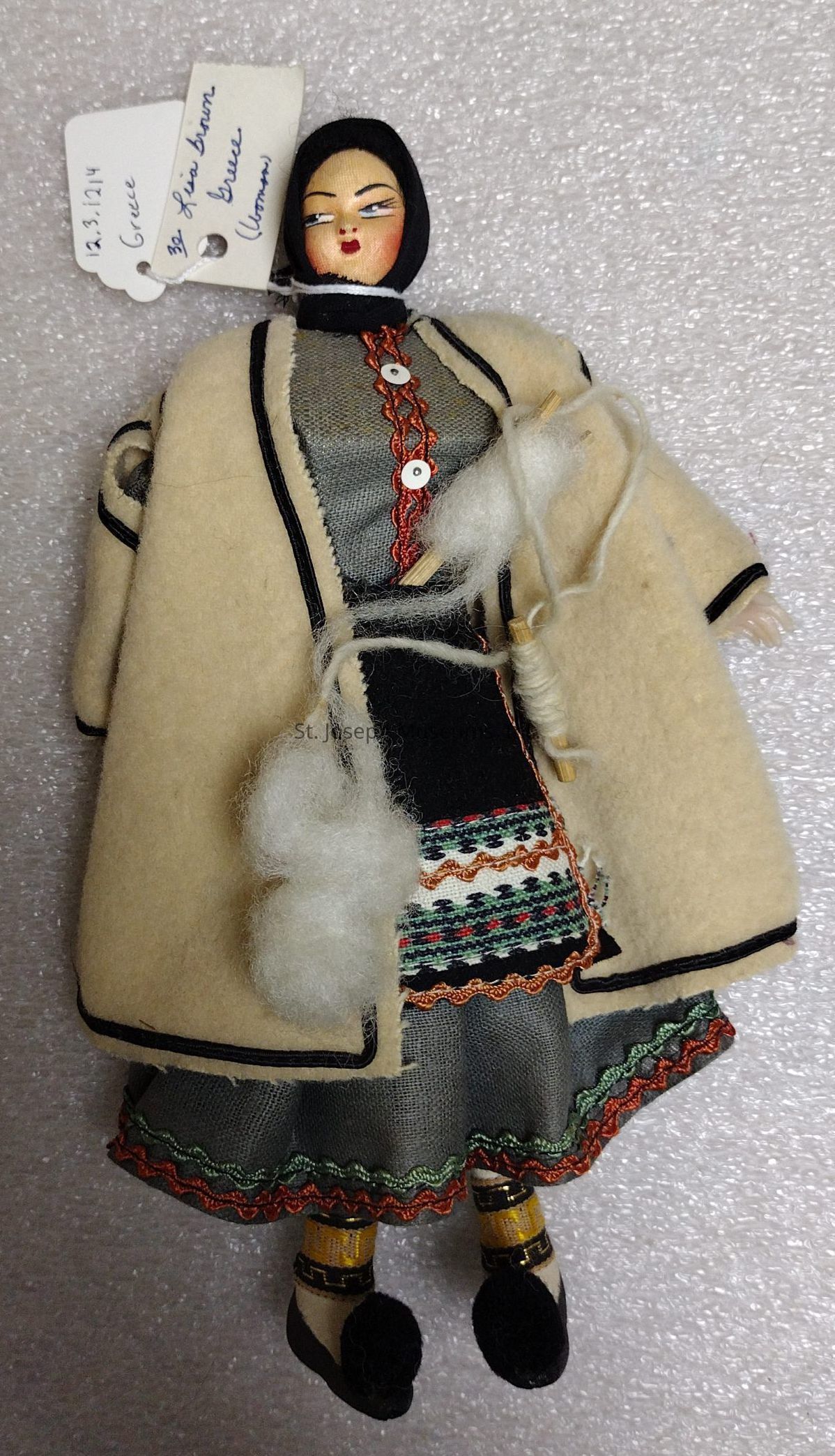 The image shows a traditional doll dressed in Greek attire, featuring a detailed traditional outfit that includes a patterned dress with colorful embroidery and a beige overcoat with black trim. The doll has a painted face with an expression, and it wears a black headscarf that covers the hair. In one hand, the doll is holding a small spindle with white wool attached, suggesting it's engaged in spinning. Attached to the doll is a small tag that appears to have handwritten notes, including references to Greece and some numbers that could indicate cataloging information, likely for a collection or exhibition.