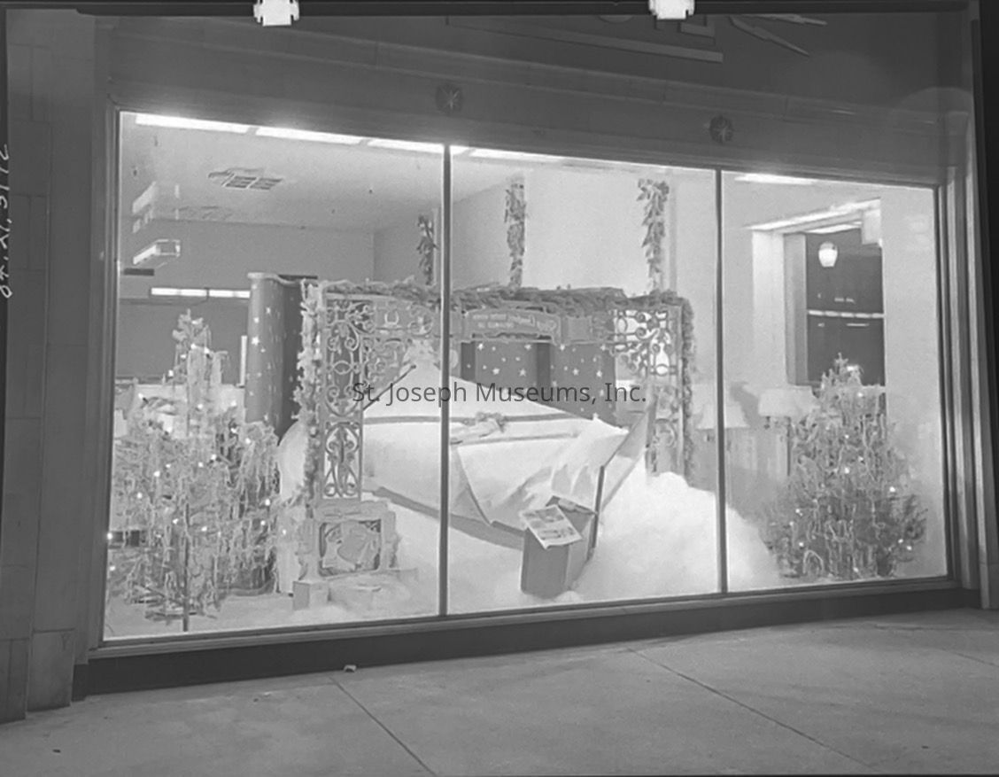 Black-and-white photograph from St. Joseph Light and Power Company of a window display at 6th & Francis Street, featuring an elegant Christmas scene with a beautifully decorated four-poster bed, surrounded by snow-like white fabric and garlands. Festive trees adorned with lights and ornaments enhance the holiday ambiance.