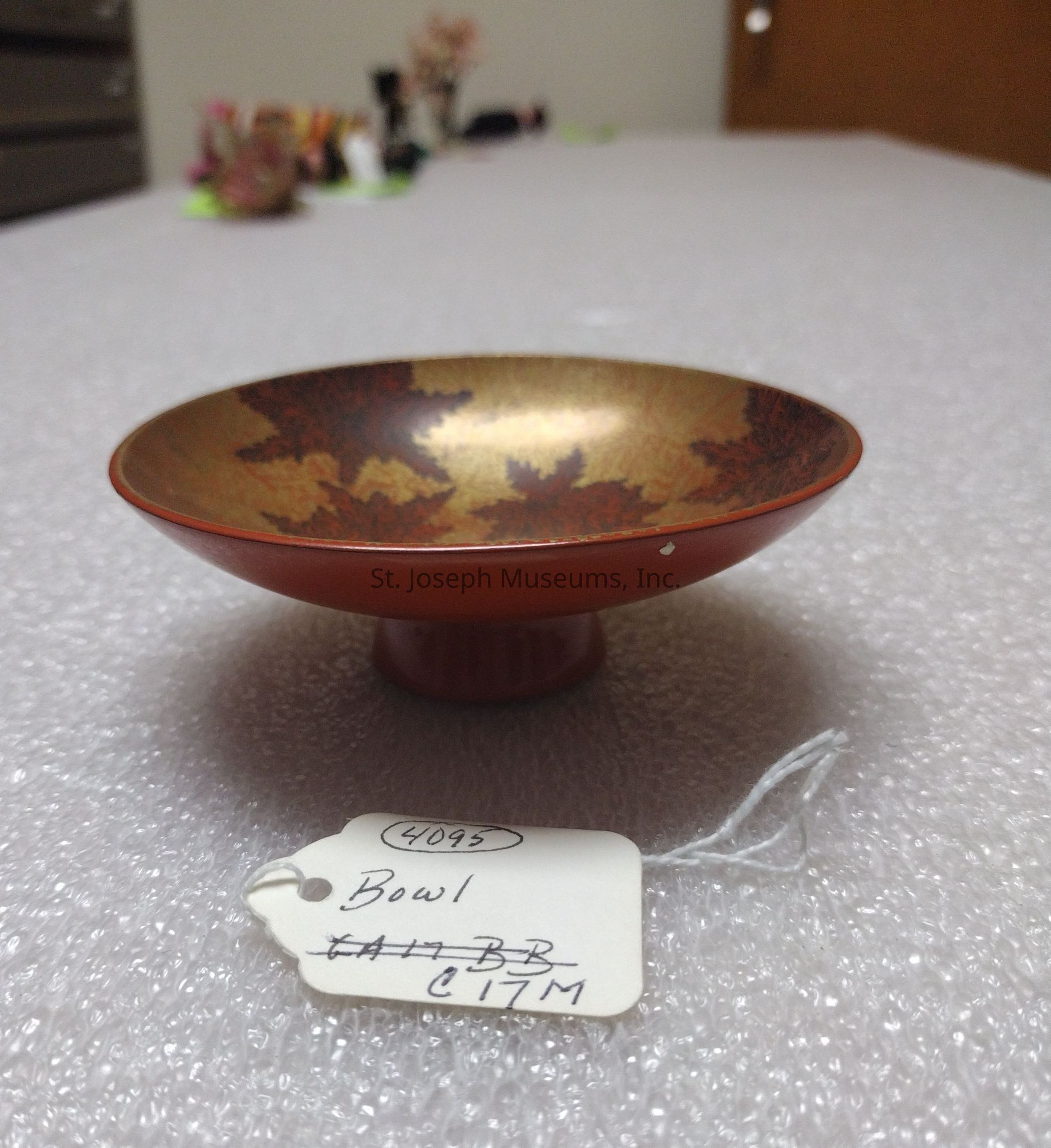 The image showcases a decorative bowl resting on a textured surface, possibly bubble wrap, displaying an intricate design featuring stylized, dark leaves on a golden background. This bowl, which appears to have a red base and a slightly flared rim, sits atop a short pedestal, giving it an elevated stance. Attached to the bowl is a string with a white tag that reads "Bowl" and has some numbers and letters on it—likely serving as an inventory or catalog reference. In the blurred background, there are various items on a table, but they're indistinct, possibly suggesting a storage or display area. The overall atmosphere hints at a setting where items might be organized or prepared, emphasizing the bowl's artistic qualities against a simple, utilitarian backdrop.