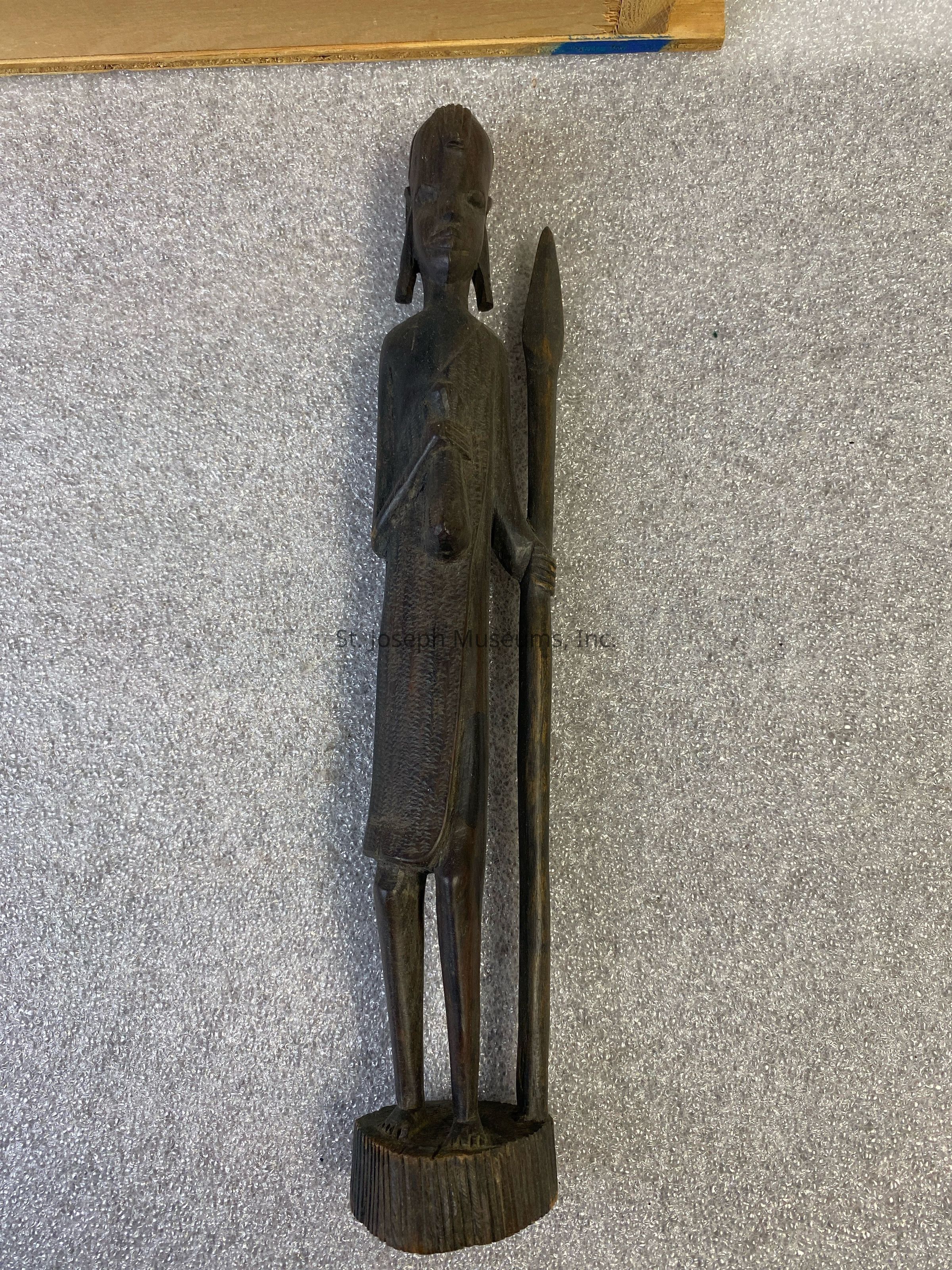 The image shows a wooden sculpture of a human figure, standing upright on a rounded base, with a detailed, textured surface. The figure appears to be wearing a garment and is holding a long spear-like object in its right hand. The sculpture has an elongated form with refined facial features, and it appears to be carved from a dark type of wood that gives it a rich, earthy appearance.