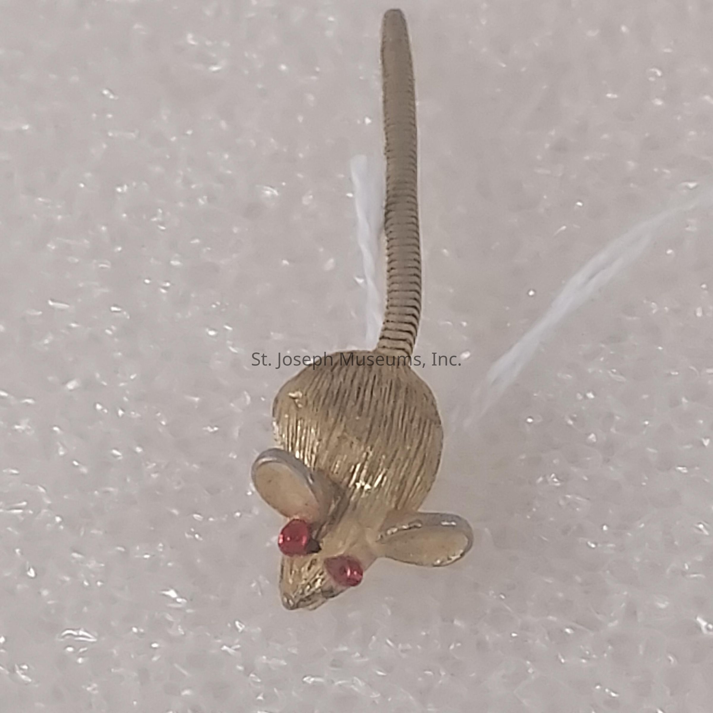 The image shows a small, detailed sculpture of a mouse, seemingly crafted from metal, with a texture that resembles fur. It has two bright red eyes that stand out prominently against its golden body. The mouse features large, rounded ears and a long, slender tail that curves slightly, adding to its lifelike appearance. The tail appears to have a spring-like texture, which adds a sense of realism and movement. This sculpture is placed on a surface that seems to be a white, textured material, possibly foam, creating a subtle contrast with the shiny metallic finish of the mouse. The lighting highlights the contours and details of the sculpture, casting soft shadows and enhancing the overall dimensionality. The craftsmanship suggests a high level of artistry, making the mouse an interesting subject of intricate design, ideal for decorative or artistic appreciation. The red eyes bring focus to the face of the mouse, adding a touch of vibrancy and character to the otherwise monochrome piece.
