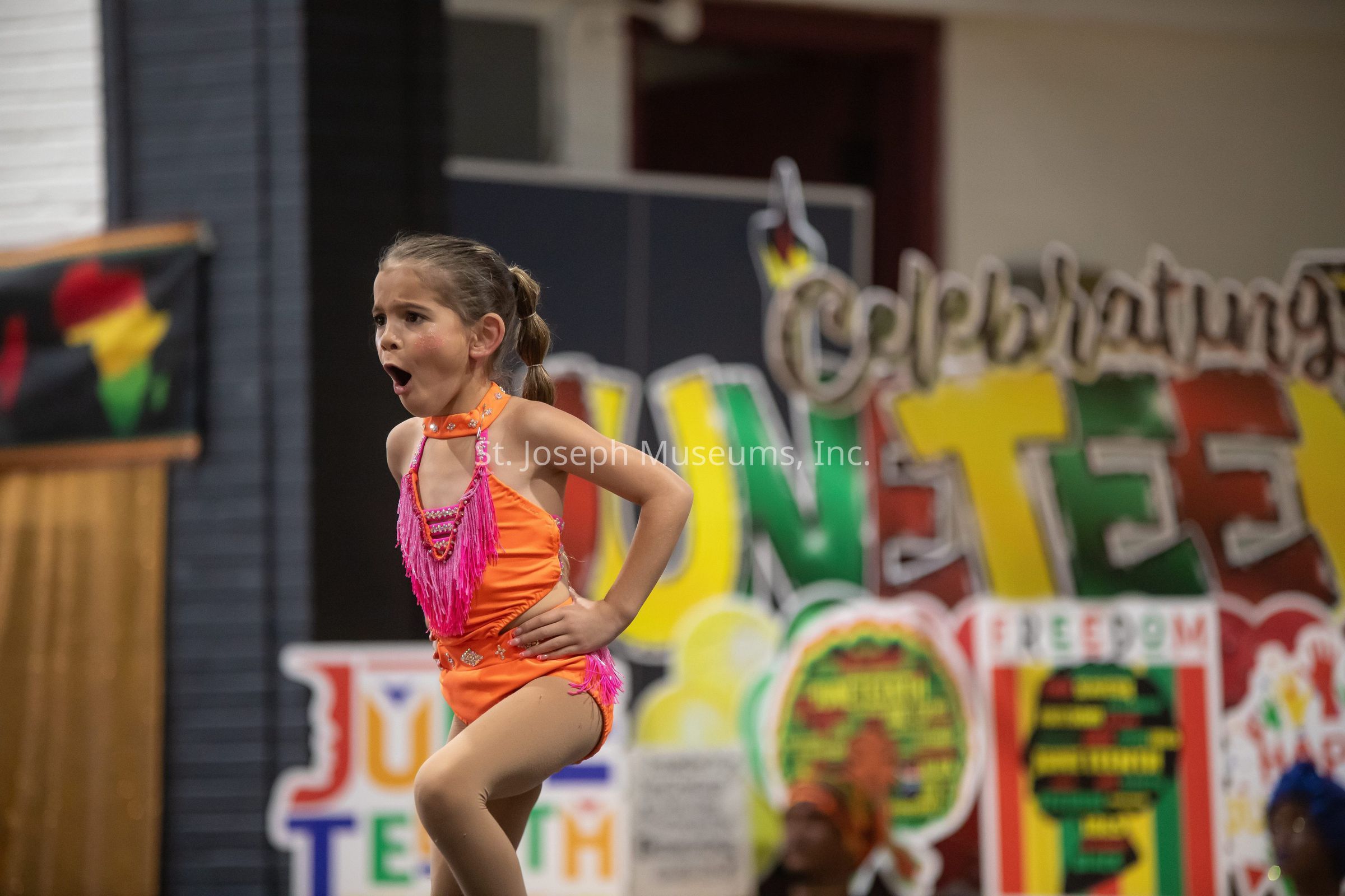 The image captures a young girl energetically performing in front of a colorful backdrop celebrating Juneteenth. She is dressed in a bright orange costume adorned with pink fringe details, and her expression shows focus and excitement. Her hair is neatly tied back, and she seems to be in the middle of a dynamic dance move. The vibrant background features large, bold letters that spell "Juneteenth" in striking colors such as yellow, green, and red, which are often associated with the celebration of African American culture and heritage. The festive atmosphere is palpable, highlighting a sense of community celebration and cultural pride.