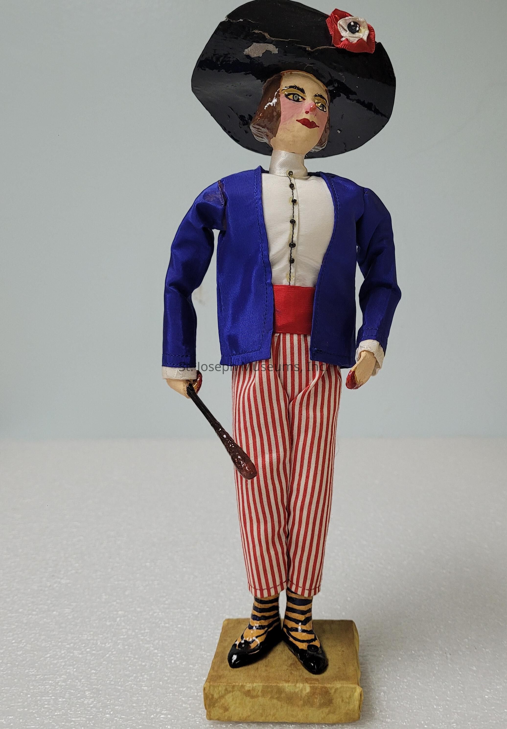 The image shows a decorative figurine standing on a rectangular wooden base, dressed in an elegant, classical outfit featuring a blue jacket over a white shirt adorned with black buttons, red and white striped pants, and a broad red waistband. The figurine's head is topped with a large, black, wide-brimmed hat decorated with a single artificial red flower. The face is painted with prominent features including rosy cheeks and a red smile. In the right hand, the figure holds a slender, dark brown baton, adding to the sophisticated appearance. The figurine’s shoes are black with a glossy finish, complemented by yellow and black striped socks, enhancing the overall whimsical yet elegant style. The background is a simple off-white surface, highlighting the figurine's vivid colors and intricate details.