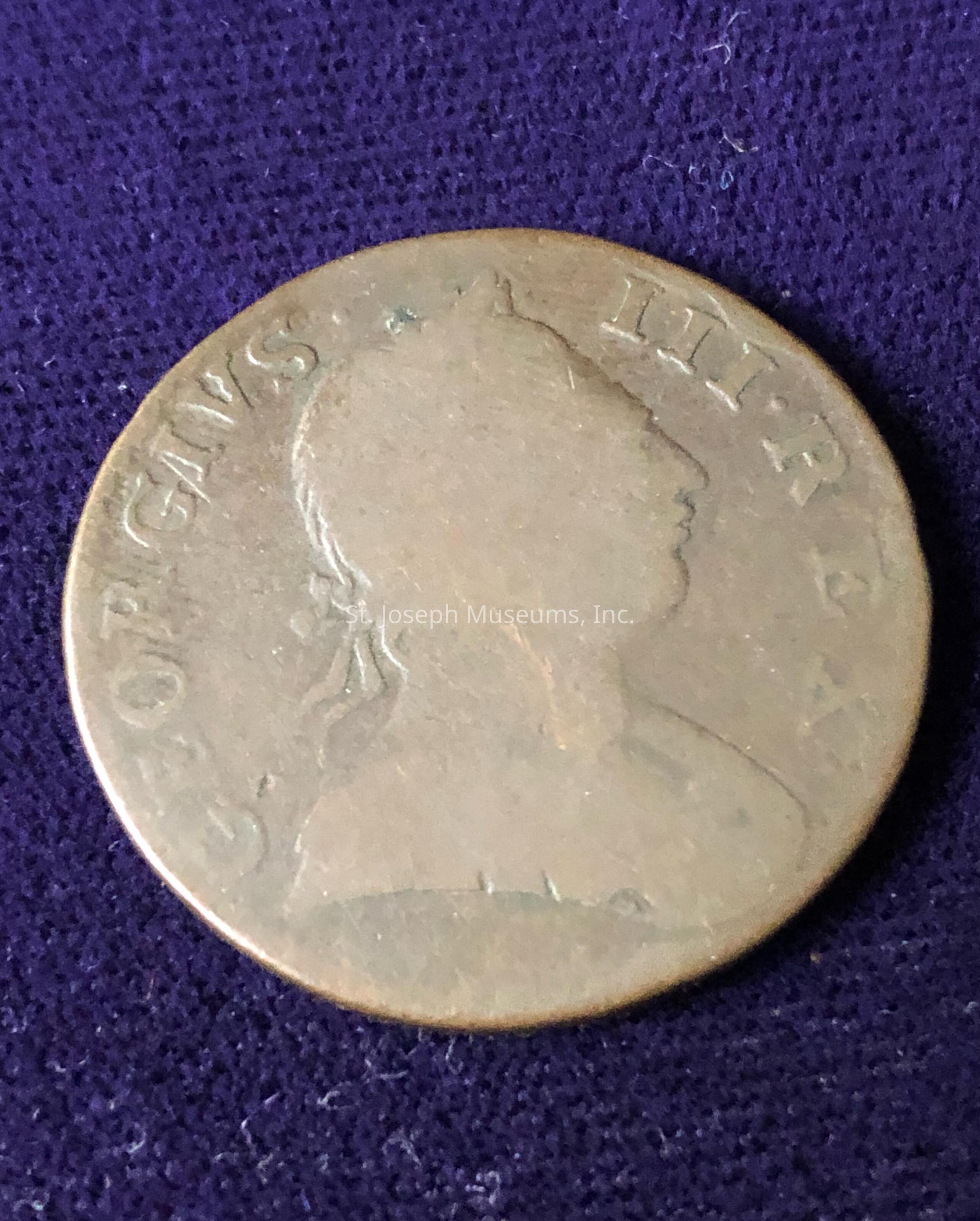 The image depicts a vintage coin with a side profile of a person's head engraved on it, surrounded by worn and partially obscured inscriptions that seem to include the letters "GEORGIVS III REX." The coin rests against a deep purple fabric background, adding contrast to the aged, brownish metal surface of the coin. The wear and patina on the coin suggest it has aged, likely indicating its historical significance or use over many years. The design and inscription hint at an association with an English monarch, possibly from the late 18th to early 19th century, commonly depicted on currency from that era.