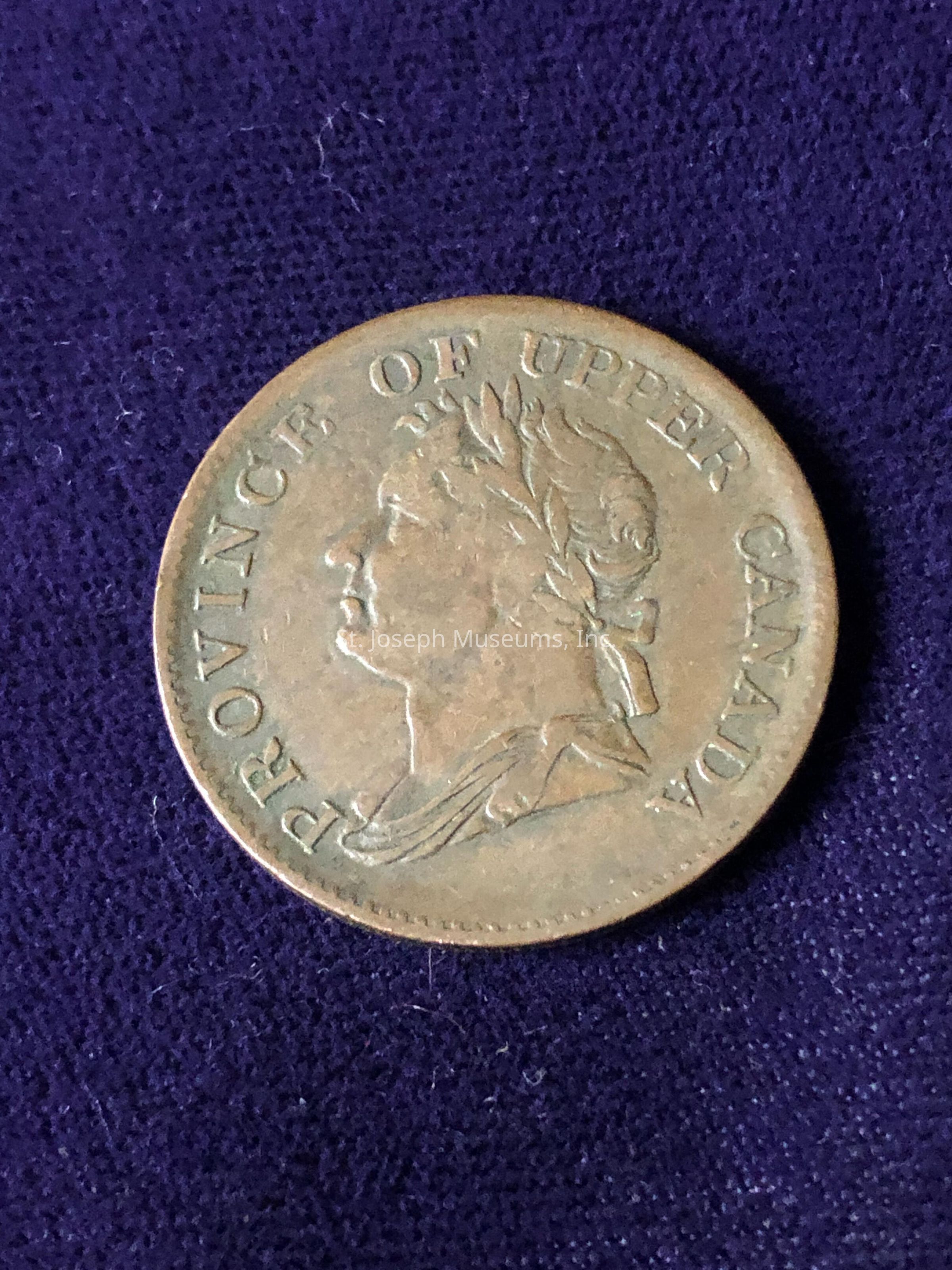 The coin features an engraving of a person's profile, adorned with what appears to be a laurel wreath, with visible text around the edge that reads "PROVINCE OF UPPER CANADA." The coin's surface shows signs of age, characterized by its patina and weathered appearance, which likely indicates its historical significance and value as a collectible item. The intricate details of the laurel and the portrait's features are delicately etched, highlighting the craftsmanship of the time.
