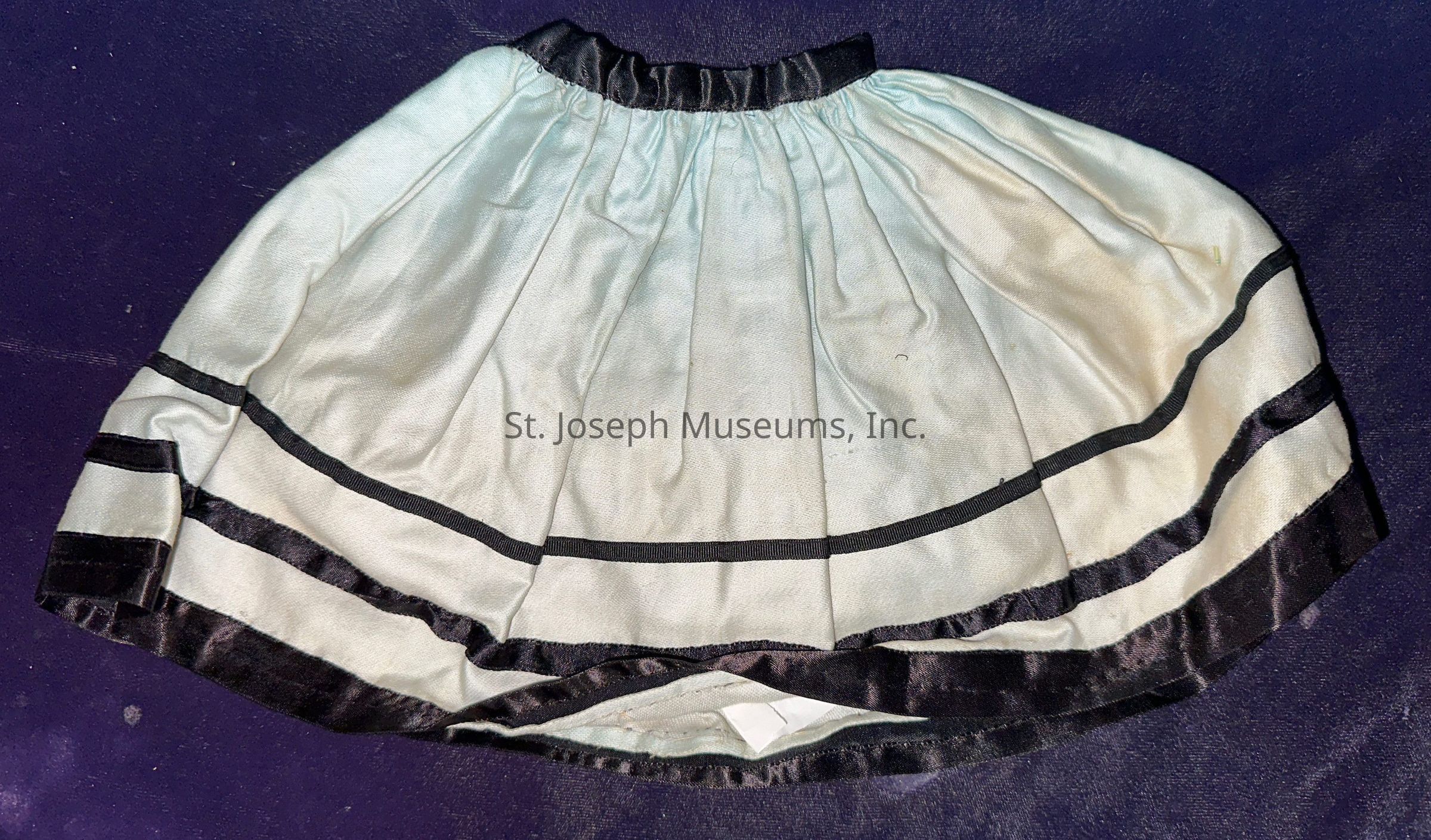 The image depicts a small doll skirt with a light fabric featuring three horizontal black velvet bands near the bottom. The skirt is gathered at the waistband, which is also made of black velvet, creating a ruffled effect. The background appears to be a dark purple surface, providing contrast to the skirt's lighter color, which enhances its visible details. The overall design suggests a classic, elegant style, suitable for a vintage or formal doll ensemble.
