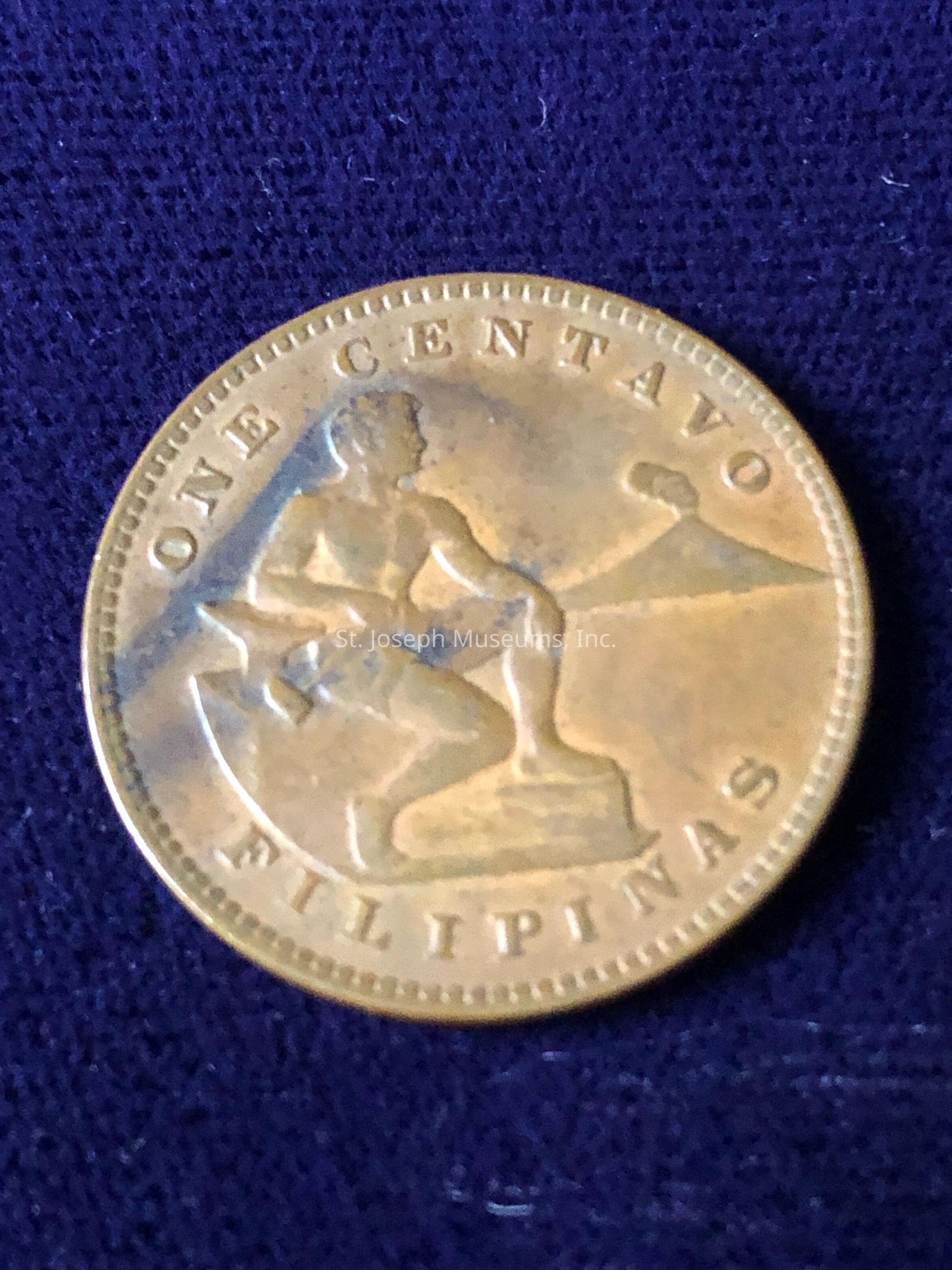 The image shows an old one centavo coin from the Philippines, featuring the inscription "ONE CENTAVO" along the upper curve and "FILIPINAS" at the bottom. The central design depicts a man seated beside an anvil, holding a hammer, with a volcano visible in the background. The coin has a bronze-like appearance, with signs of wear and a slightly tarnished surface, suggesting age. The details are intricate, highlighting the craftsmanship involved in its creation.