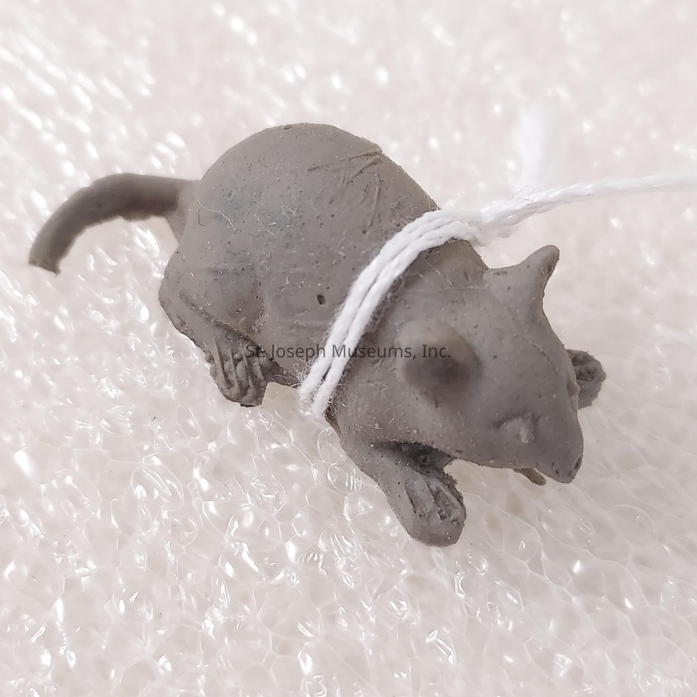 The image depicts a small, gray, sculpted figure of a mouse, lying on its stomach with its legs bent under its body. The mouse sculpture has a realistic appearance, capturing details such as its small ears, closed eyes, and long tail. A white string is tied around the middle of the sculpture, securing it against a textured, white surface that resembles bubble wrap. The overall impression suggests a meticulously crafted miniature with an emphasis on detail, contrasting the grayness of the mouse with the bright, textured background, offering a sense of fragility and artistic expression.