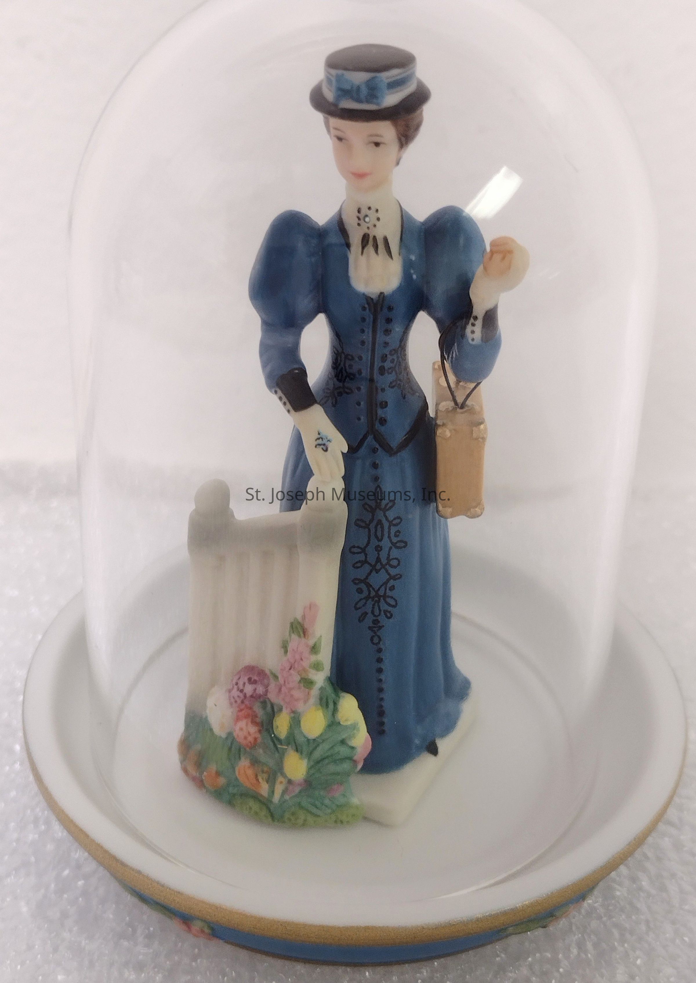 The image shows a delicately crafted porcelain figurine encased in a clear glass dome, depicting a woman dressed in an elegant blue Victorian-era dress adorned with intricate black patterns and puffed sleeves, accentuated by a matching hat with a blue ribbon. She is holding a brown handbag in one hand and resting the other on a white fence adorned with vibrant, colorful flowers at its base. The entire ensemble rests on a white base with a gold rim, offering a charming and nostalgic representation of a bygone era, capturing the grace and style of a historical figure preserved within a protective transparent covering.