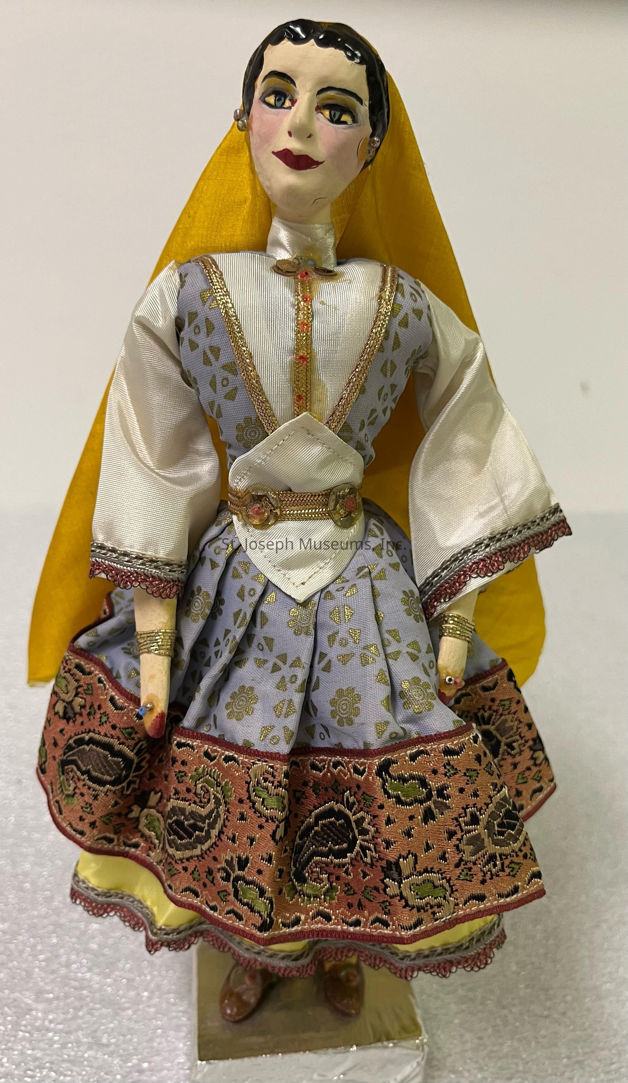 The image shows a detailed figurine of a person in traditional attire, featuring intricate craftsmanship. The figure wears a vibrant costume with a white and gray patterned dress adorned with elaborate golden accents, and the sleeves are made of lustrous fabric. A yellow veil drapes from the head, adding contrast to the composition, while a decorative belt accentuates the waist, showing ornate red and gold designs. The hem of the dress is embellished with a rich, multicolored border with paisley and floral patterns. The face of the figurine is expressive, painted with bold eyebrows, rosy cheeks, and vibrant red lips. Small earrings add further detail to the facial features, and delicate jewelry is visible on the wrists. This artistic piece stands on a textured surface, balancing on what appears to be a square pedestal, creating an elegant and culturally rich presentation.
