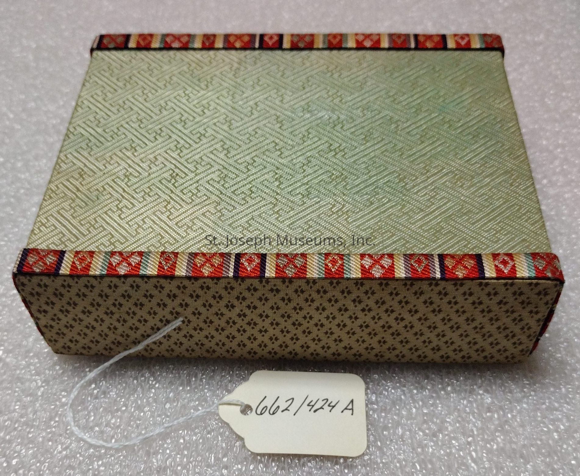 The image displays a rectangular box with a decorative design, featuring a light green lid adorned with an intricate pattern of interlocking lines. The edges are bordered by a colorful strip with alternating red and gold geometric motifs, adding a striking accent to the lid. The sides of the box have a subtle pattern consisting of small, dark shapes on a light background. Attached to the box is a white tag with the handwritten number "662/424 A." The surface on which the box rests is textured, appearing to be a soft material that cushions the box. The overall appearance suggests a blend of traditional and artistic craftsmanship.