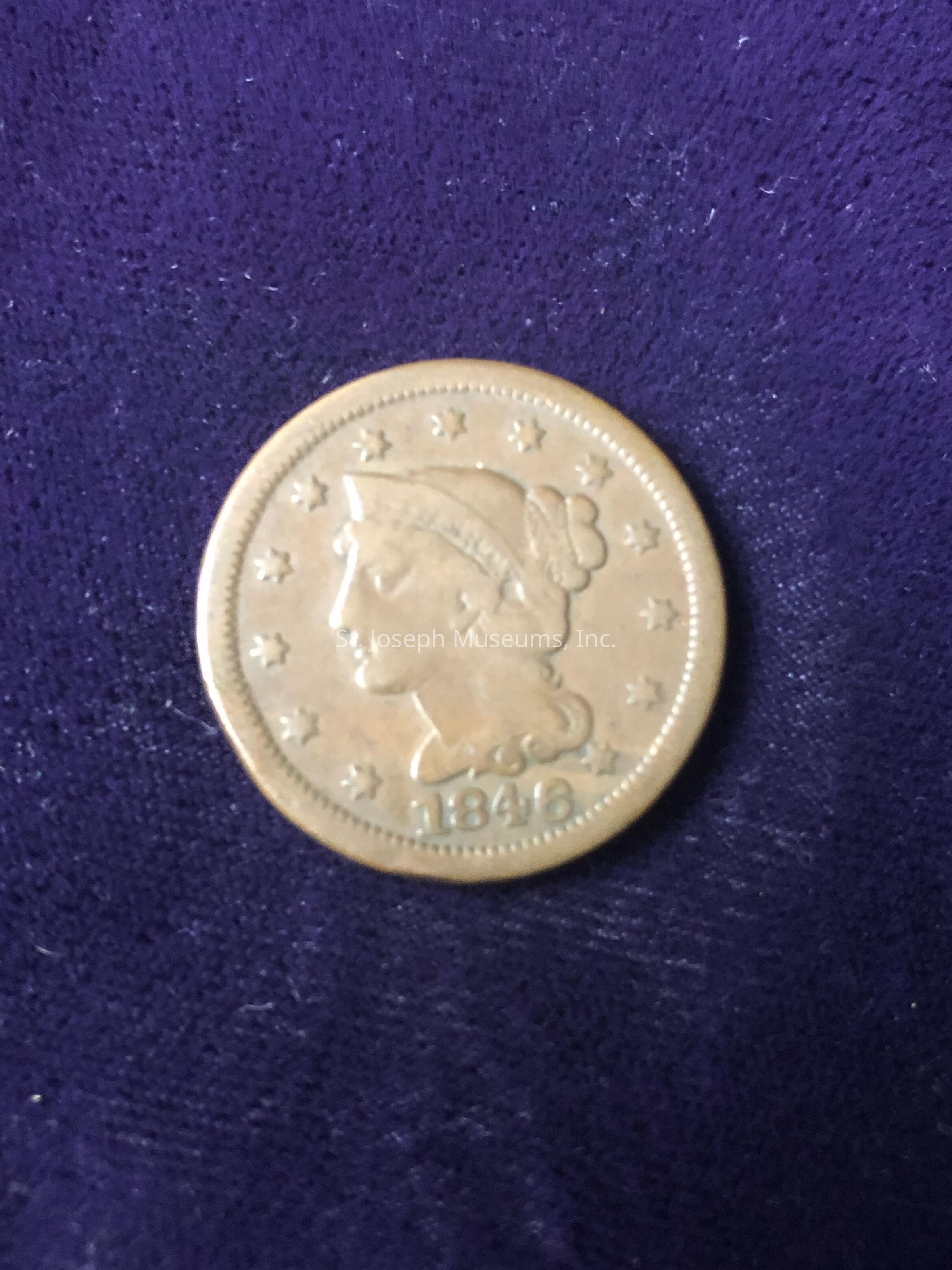 Featuring a profile of a woman's head in the center with the word "LIBERTY" inscribed on her headband, surrounded by thirteen stars symbolizing the original American colonies. This coin is dated 1846 at the bottom, indicating it was minted in the mid-19th century. The obverse design indicates it is likely a U.S. Liberty Head coin, possibly made of gold, considering the historical context. The intricate details of the design, such as the woman’s hairstyle and the stars, reflect the artistic style of the period, making this coin not only a piece of currency but also a piece of history that offers insight into the era's numismatic design. The coin appears to be well-preserved, suggesting it has been carefully maintained by collectors or historians. Its presence on the rich purple fabric enhances the antique and collectible nature of the item, suggesting it could be a prized piece in a collection, representing the United States