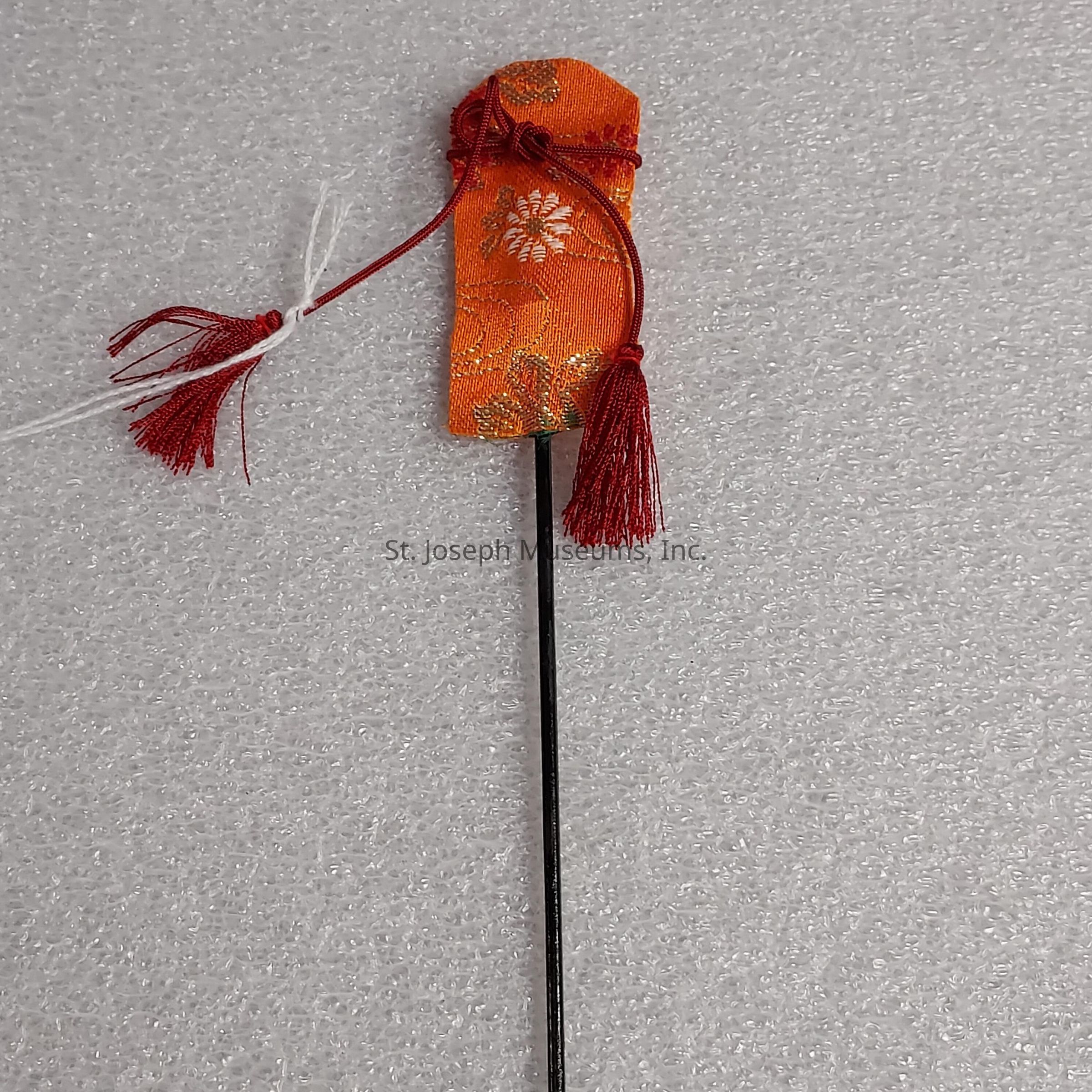The image shows a decorative hairpin, featuring a long, slender black stick, topped with an intricately designed fabric ornament. The fabric is vibrant orange, adorned with floral patterns in shades of gold and white, adding a touch of elegance. Attached to the ornament are red and white threads, tied in a loop, with red tassels cascading down, providing a striking contrast and enhancing the aesthetic appeal. The backdrop is a simple, textured white surface, which accentuates the vivid colors and delicate craftsmanship of the hairpin, highlighting its cultural and artistic significance as an accessory.