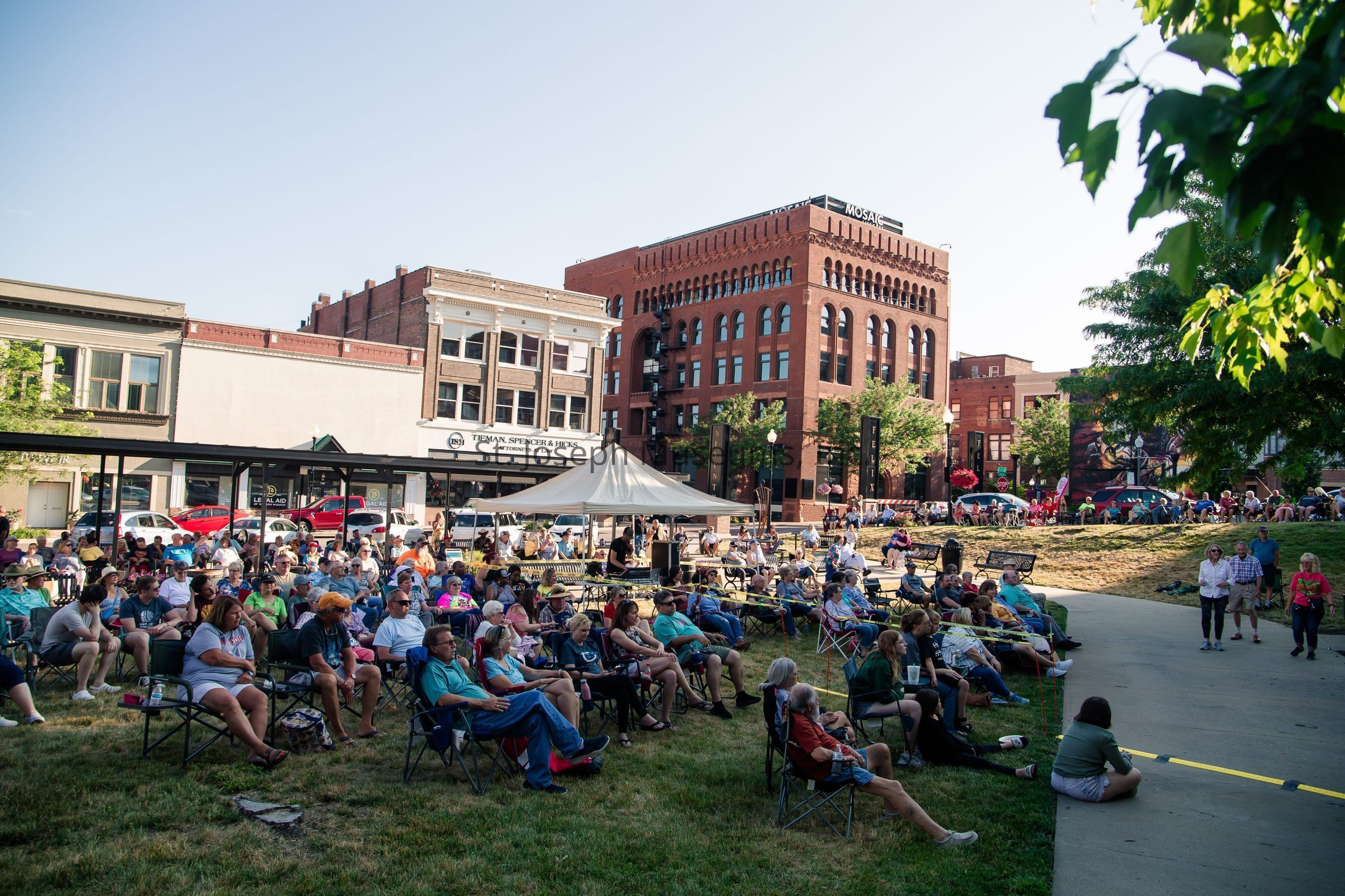 The image depicts a lively outdoor gathering with a large crowd of people seated on foldable chairs and the grass, all facing a central area that appears to be the focus of the event; this gathering takes place in a park-like setting surrounded by historic brick buildings and a few modern commercial establishments, indicating an urban downtown area. The sky is clear and blue, casting a warm, sunny ambiance over the scene. A white tent, possibly housing event staff or equipment, is set up in the middle, while cars can be seen parked along the street. People of various ages are enjoying the event, with some individuals standing under the shade of nearby trees, adding to the relaxed and casual vibe of the gathering. The architecture of the surrounding buildings features large windows and ornate brickwork, contrasting with the greenery of the park.