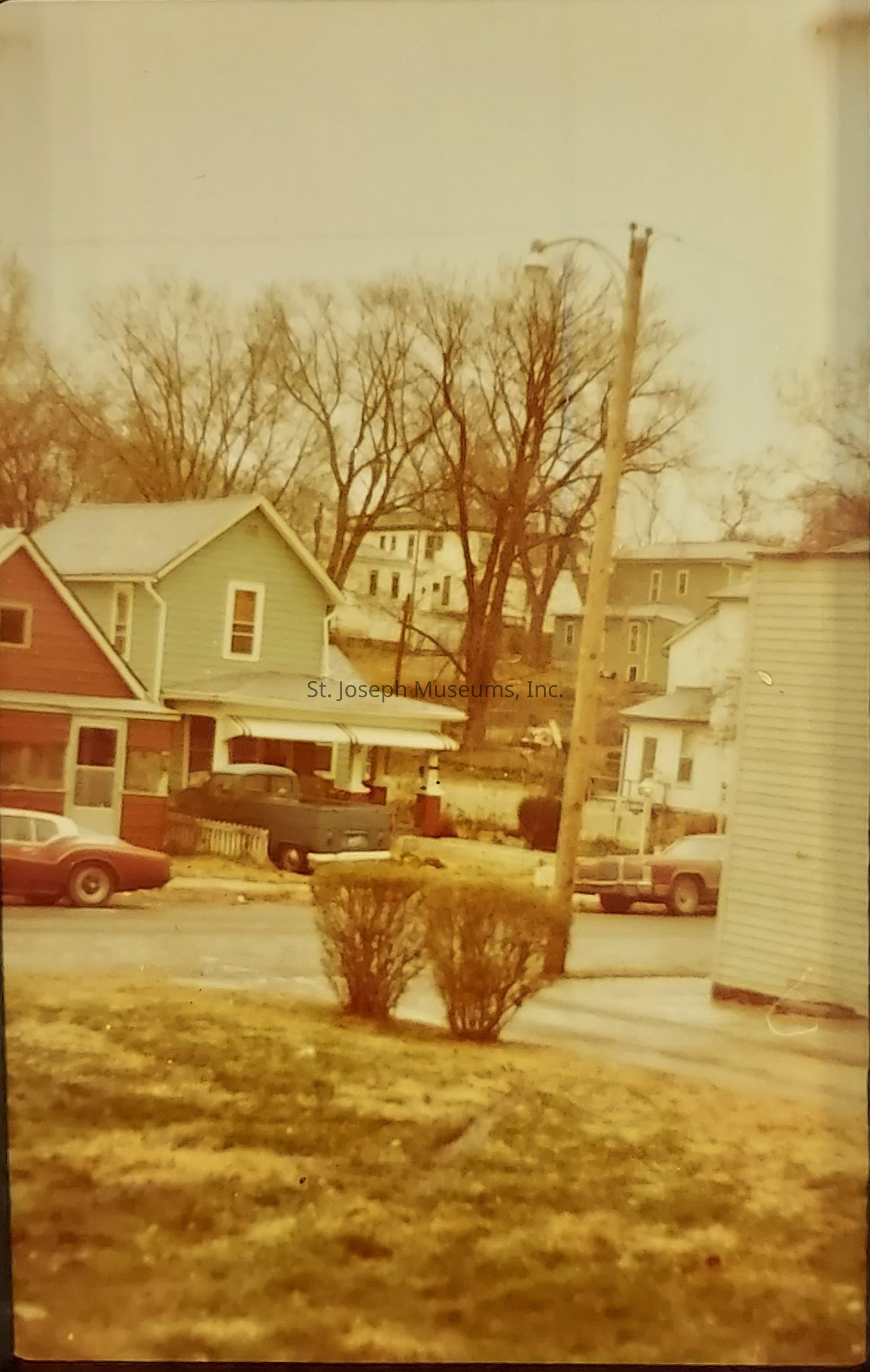 The image depicts a quaint, vintage suburban scene characterized by a blend of homes with a very retro appearance, possibly from the late 20th century, showcasing several houses lined along a quiet street. In the foreground, a classic car and a pickup truck are parked outside a two-story house painted in a muted green hue, accompanied by another house with a red-brown exterior. Leafless trees surround the area, hinting at a winter or early spring setting. Power lines and a utility pole stretch across the scene, adding a touch of everyday life. The distant backdrop reveals more homes nestled in a slightly elevated area. Indistinct, bare bushes and a patchy, grassy yard are visible, capturing the essence of a quiet, nostalgic neighborhood, where time seems to linger in a past era, evoking memories of simple, bygone days.