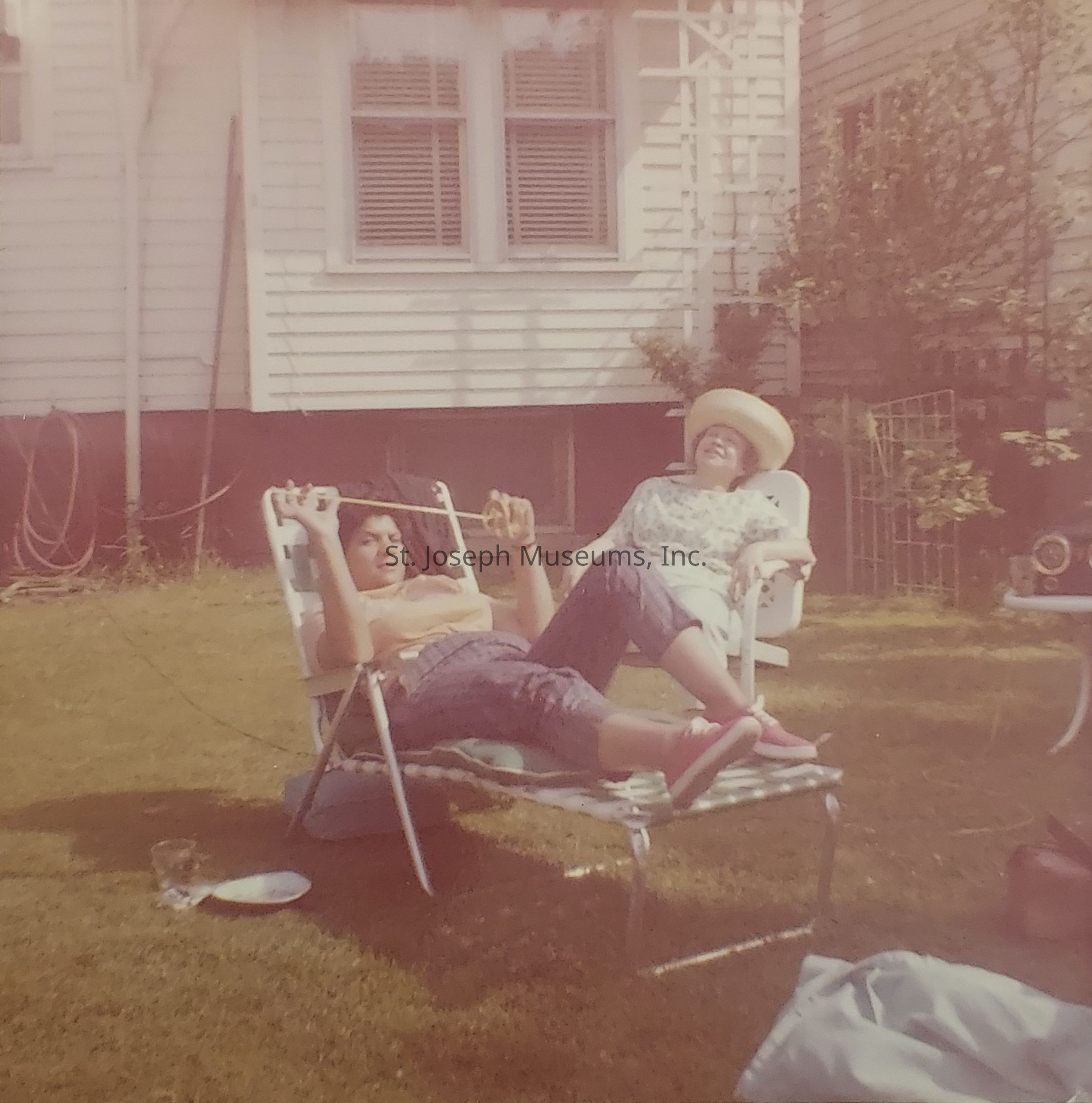 In this vintage image, two people are relaxing on lounge chairs in a sunlit backyard with a white house in the background. The person on the left is lying back with a relaxed demeanor, holding what appears to be a straw, while dressed casually. The person on the right is sitting comfortably, wearing a straw hat, and appears content. The scene captures a carefree, leisurely moment, with the grass and a coiled hose visible in the background. Nearby, there's a glass and a plate on the ground, adding to the laid-back atmosphere of the day.