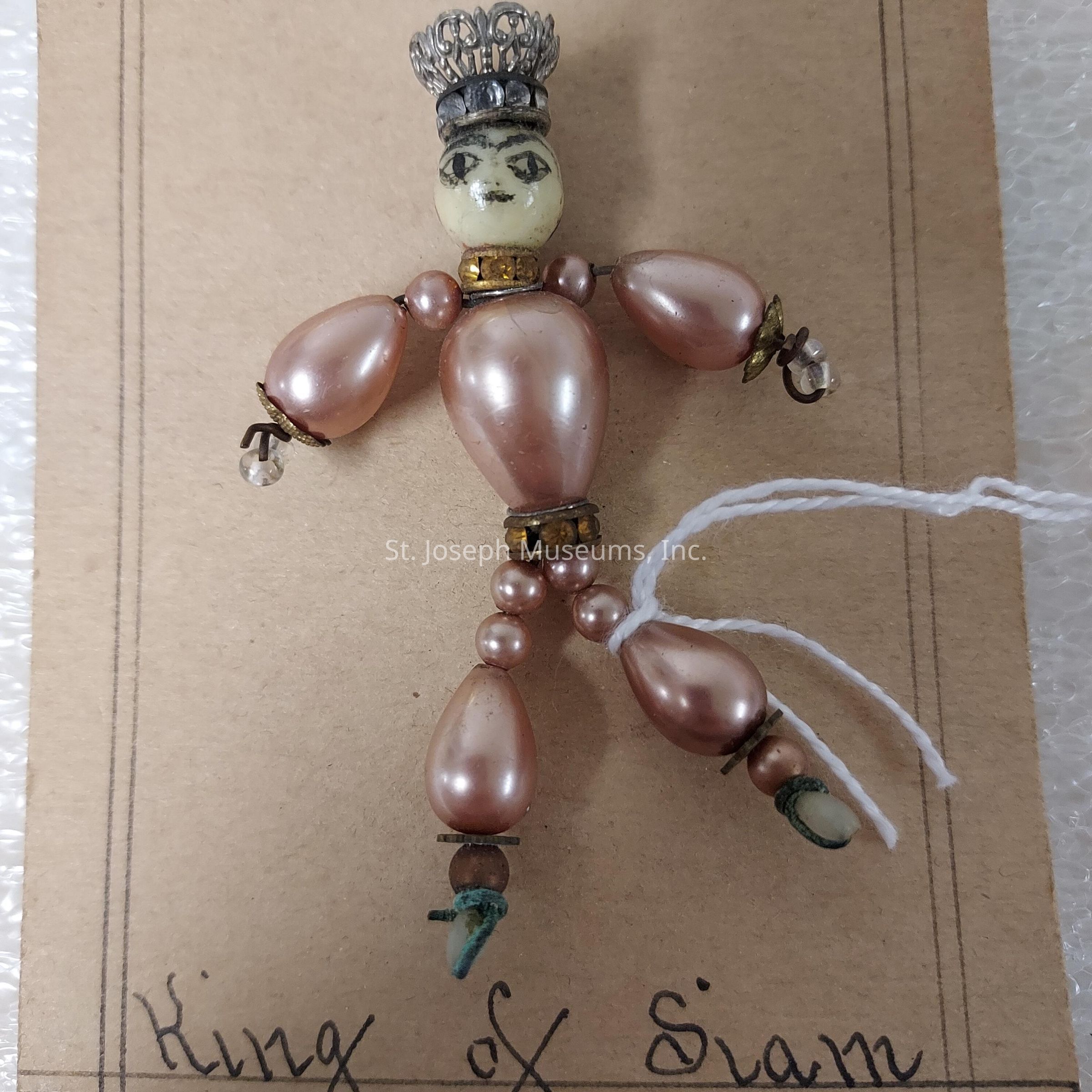 The image depicts a charming handcrafted figure labeled "King of Siam." The figure is composed primarily of pearl-like beads, with larger beads used for the torso and limbs, giving it a doll-like appearance. It features a small, intricately designed silver crown perched on its bead head, which is painted with delicate facial features, including expressive eyes and a subtle mouth. The arms and legs are articulated with smaller beads acting as joints, allowing for a flexible pose. A piece of white string is attached to one leg, possibly for hanging or positioning the figure. The background consists of a simple piece of cardboard with handwritten text identifying the figure as the "King of Siam," providing a touch of personalization and historical reference.