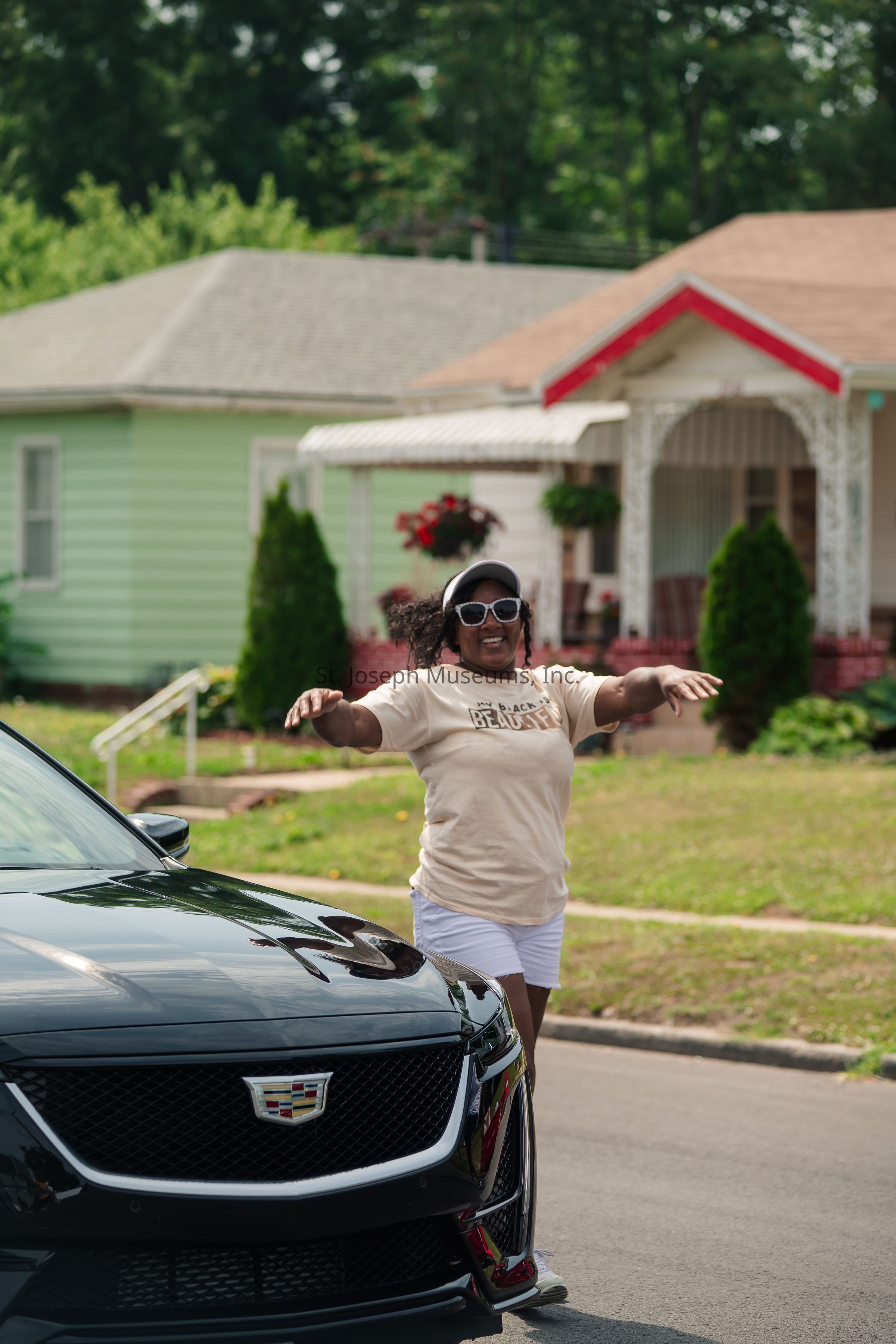 In the image, a woman is joyfully standing with her arms outstretched in front of a sleek black Cadillac parked on a suburban street. She is dressed casually in a beige t-shirt with a positive message, white shorts, and sneakers, while also sporting sunglasses and a cap. The background reveals a charming neighborhood with well-maintained lawns and houses adorned with vibrant painted exteriors. The house directly behind her features a pastel green color with a tree and shrubbery, while another nearby house is predominantly white with red accents and lush greenery. The scene conveys a cheerful and relaxed suburban atmosphere.