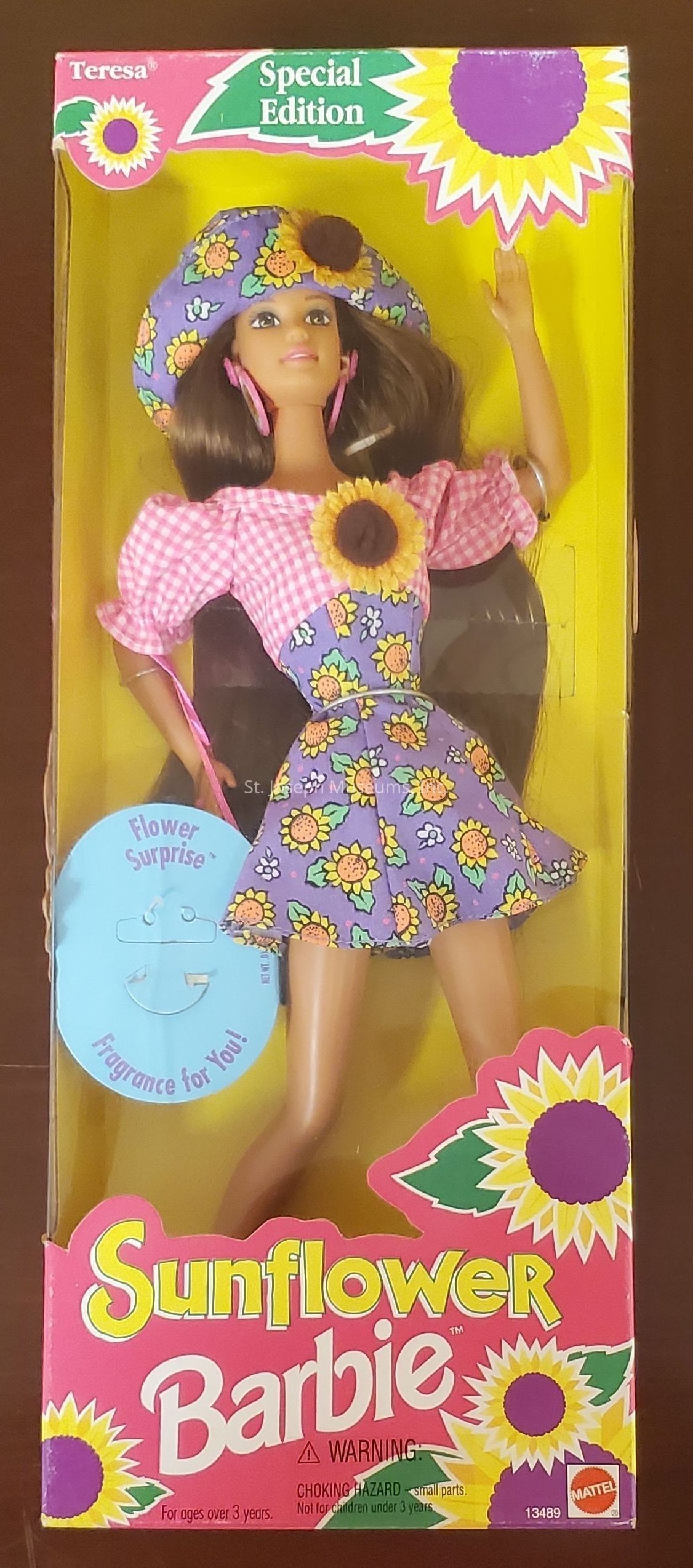 The image depicts a “Special Edition” Sunflower Barbie doll in its original packaging. The doll is designed with a charming sunflower theme, featuring a vibrant outfit adorned with sunflower patterns on a purple dress and matching hat, accompanied by a pink checkered blouse. The packaging is colorful, with a predominantly pink background and multiple sunflower graphics, reflecting the doll’s theme. The box also highlights a “Flower Surprise” fragrance feature for the owner. The text "Teresa" is seen at the top left, indicating a character name, while the iconic Barbie logo is prominently displayed at the bottom, along with a warning about small parts and a reminder for ages three and up.