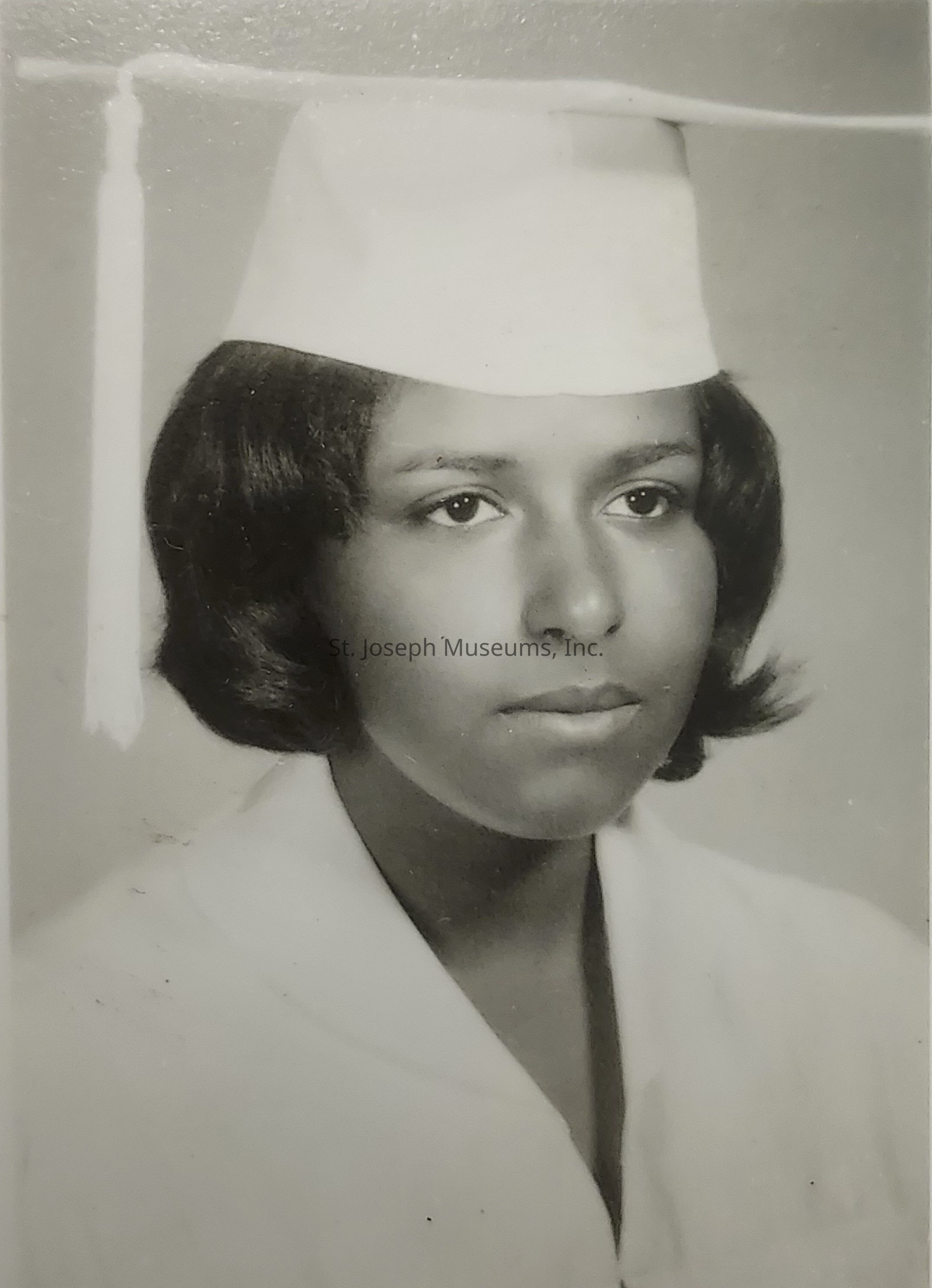 The black-and-white photograph portrays a young woman in a graduation cap and gown, her expression composed and thoughtful, as she gazes slightly away from the camera; her graduation attire consists of a neatly worn white cap, complete with a tassel, and a matching gown, symbolizing a significant milestone in her educational journey, while her short, well-kept hair frames her face in soft waves, adding an elegant touch to the formal portrait, and the monochrome setting emphasizes the timelessness of the moment captured, suggesting the pride and accomplishment that accompanies a graduation, as well as the anticipation of embarking on a new chapter of life, embodying both the culmination of past efforts and the promise of future possibilities.