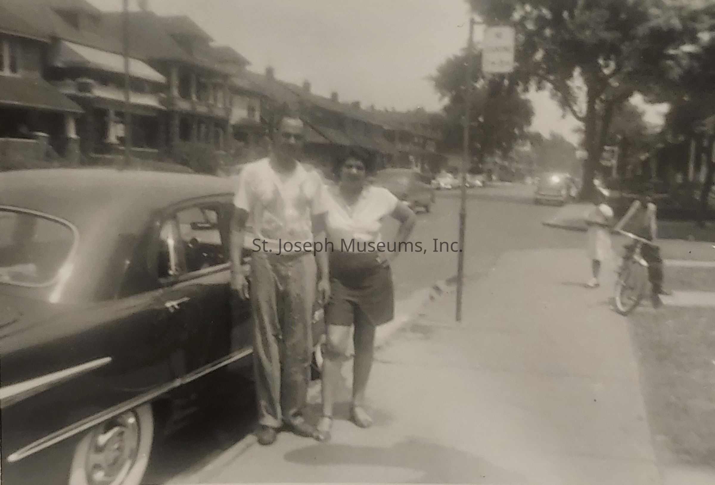 In this vintage, black-and-white photograph, a man and a woman stand closely together on the sidewalk beside a classic car, suggesting a scene from the mid-20th century. The street is lined with similar-looking row houses, hinting at a residential neighborhood, while the absence of color evokes nostalgia and a sense of history. The man is dressed in casual attire featuring a t-shirt and trousers, and the woman wears a blouse and shorts, both embodying a relaxed mood typical of a summer day. Nearby, a child with a bicycle is visible, adding a touch of daily life to the setting, with trees lining the street enhancing the pictorial depth and creating a peaceful urban backdrop.