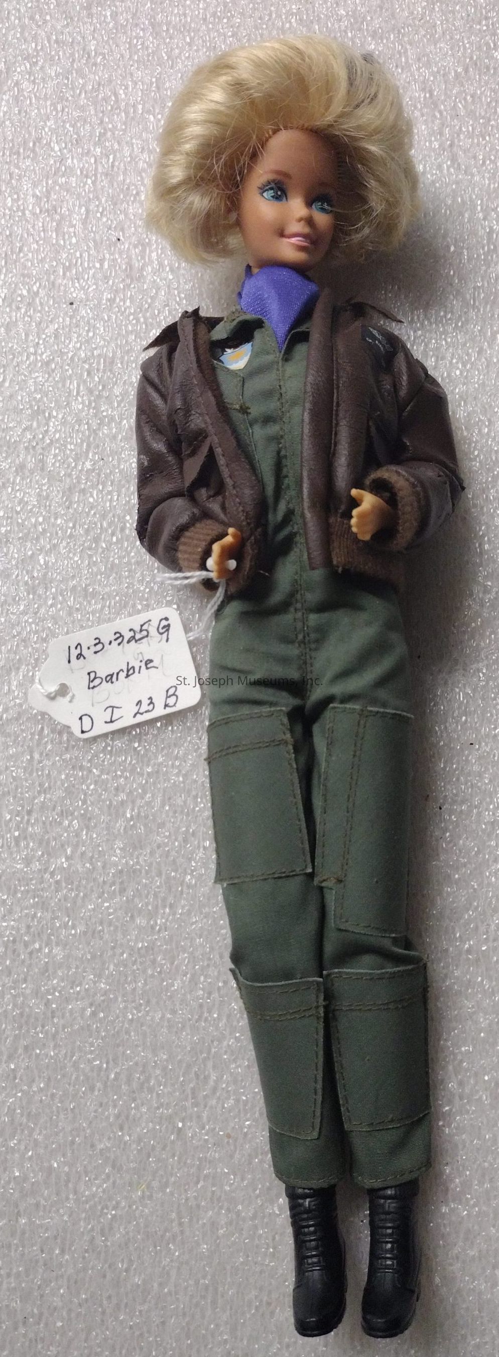 The image shows a doll with short, blonde hair, wearing a brown jacket over a green jumpsuit, and black boots. The doll features light blue eyes and a faint smile. Attached to the doll is a handwritten label reading "12.3.325 G Barbie D I 23 B," likely indicating an identification or collection number. The background is a textured, off-white surface that seems to be a type of packaging material, such as Styrofoam or bubble wrap, enhancing the perception that this doll might be part of a catalog or collection, possibly used for display, archival, or sale purposes.