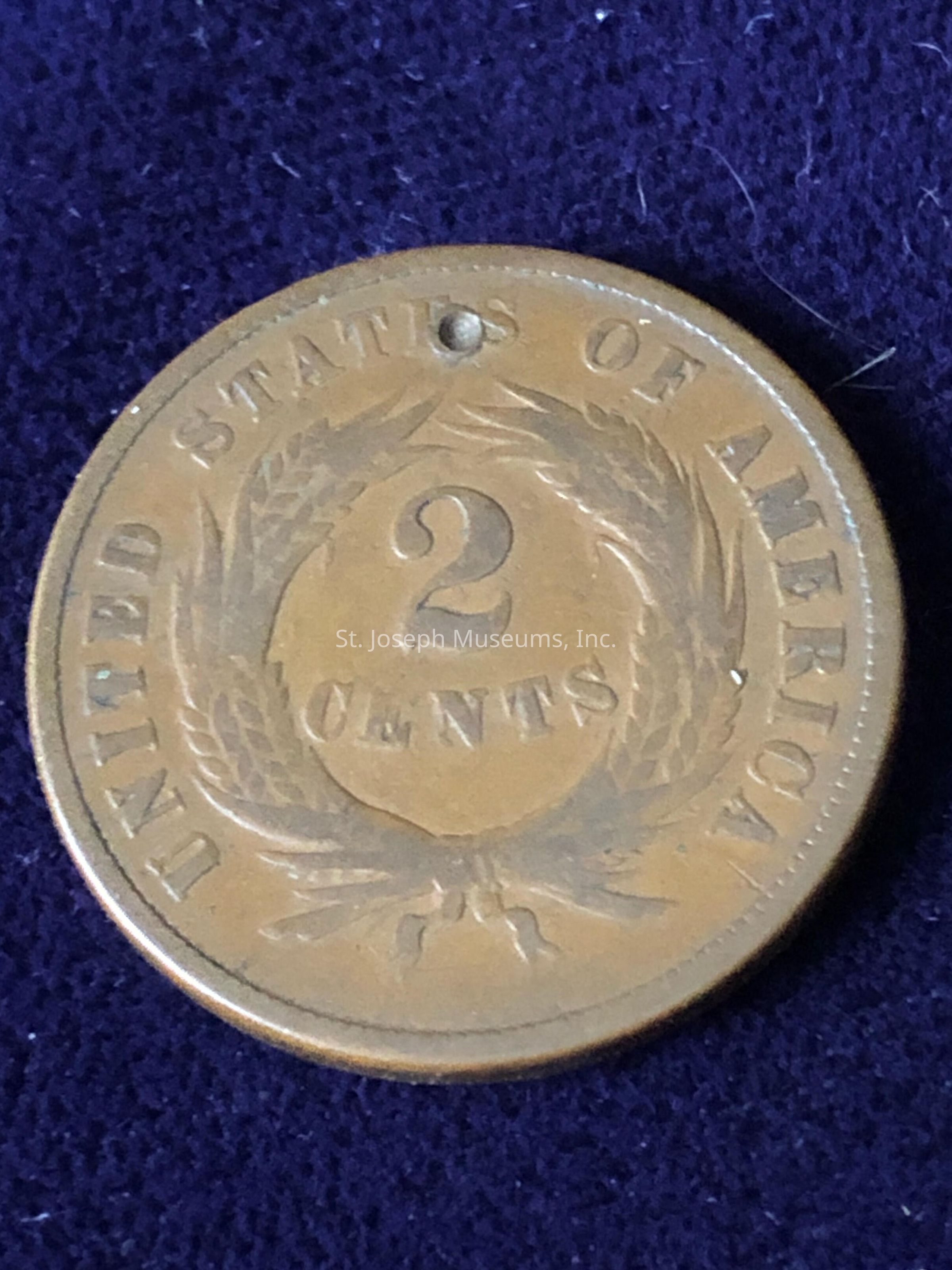 The image shows a vintage two-cent coin from the United States of America, featuring the words "UNITED STATES OF AMERICA" around the outer edge, surrounding a design with a large numeral "2" in the center, positioned above the word "CENTS" which is elegantly inscribed below. The design features a laurel wreath encircling the central text, symbolizing victory and honor, with intricate detailing that reflects the craftsmanship of the time. The coin appears to have a hole near the top, suggesting it may have been used as a pendant or simply worn over time. The coin possesses a rich, aged patina, lending it a historical appearance, while the background is a dark, textured fabric, possibly felt, which enhances the contrast and clarity of the coin's details.