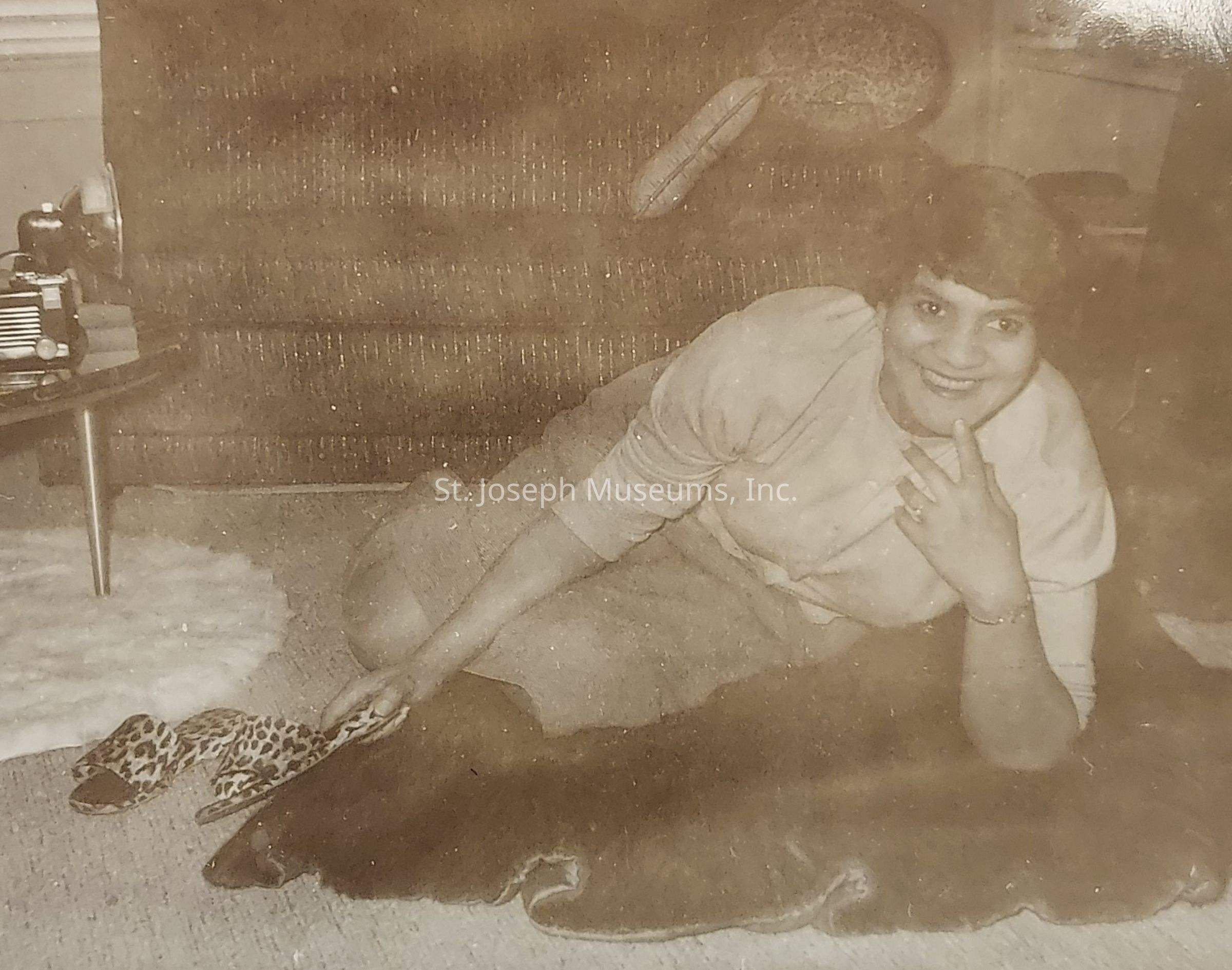 The sepia-toned image captures a woman lounging comfortably on a plush carpet, smiling warmly at the camera while resting on her side in what appears to be a cozy living room; she wears a light-colored top and skirt, and her hand gracefully supports her head, which is adorned with short hair, her other hand gently placed on the carpet, with a pair of leopard-patterned slippers visible near her feet, reflecting a relaxed and intimate moment, the background features a textured sofa with a patterned pillow resting on it, a small table stands nearby holding what looks like a vintage camera or radio, adding a mid-century ambiance to the setting, as soft natural light filters through a window, hinting at a serene evening inside a welcoming home.
