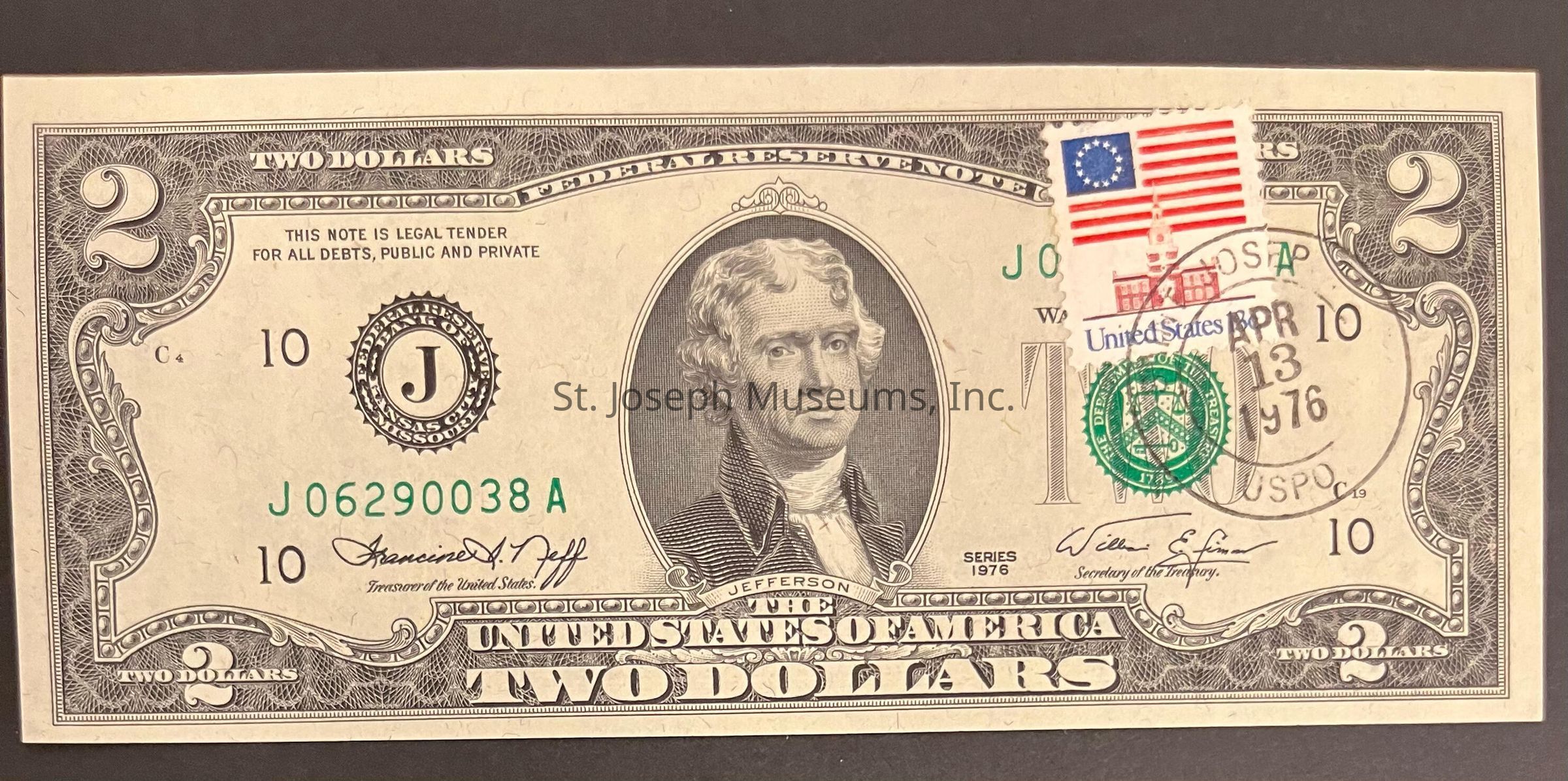 The image shows a United States two-dollar bill featuring a portrait of Thomas Jefferson, with intricate detailing on its borders and a large numeral "2" in the corners. The bill is marked as legal tender and includes signatures of government officials. Notably, there is a stamp on the right side, marked with a cancellation date of "APR 13 1976," showing a red and blue motif resembling the early American flag and a historical building. The serial number "J06290038A" is printed in green to the left side, and there are Federal Reserve and Treasury seals on either side of Jefferson's portrait, adding to the historical design elements that combine American history and finance.
