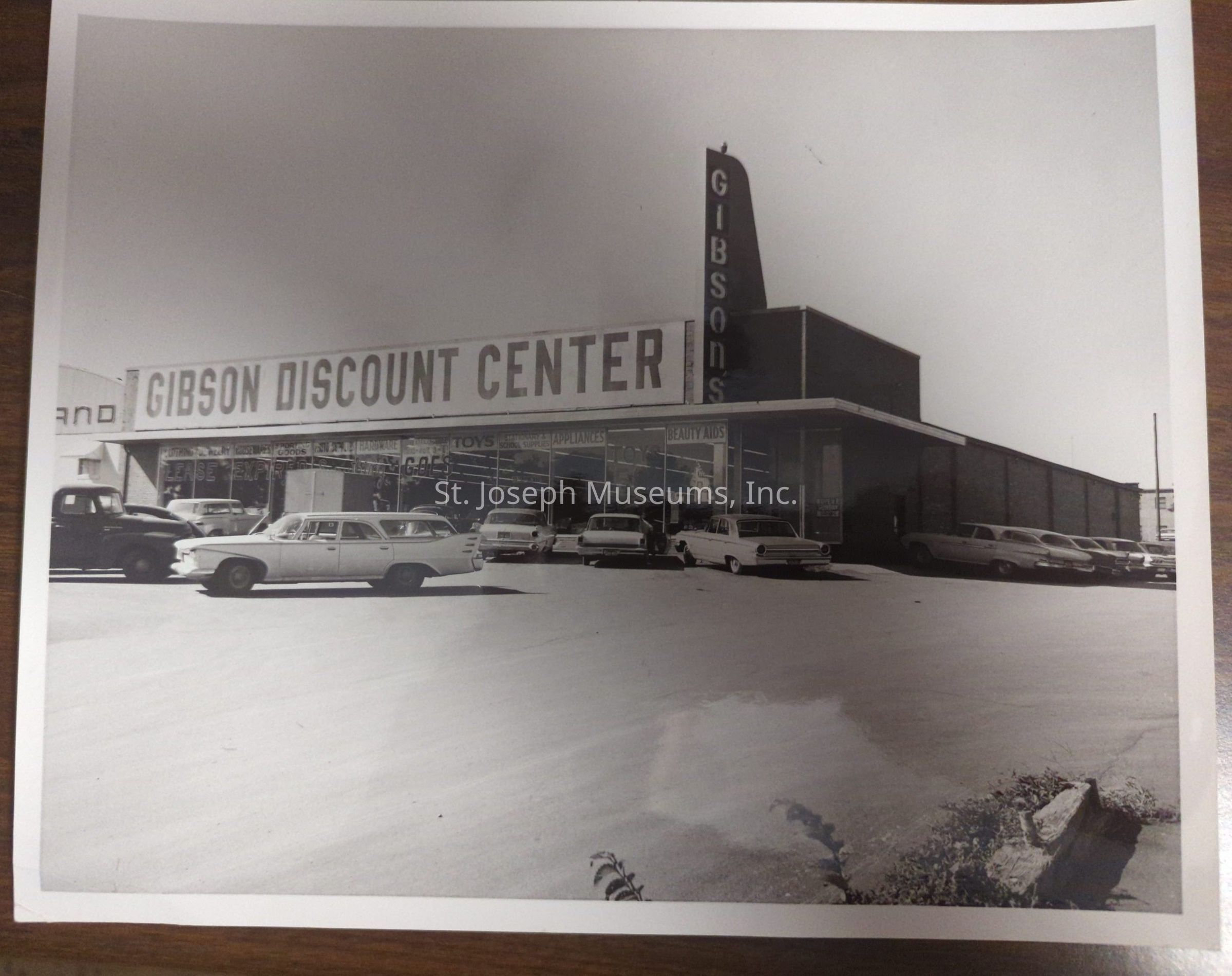 Black-and-white photo depicting the exterior of the Gibson Discount Center located at 3609 Frederick, with several classic cars parked in front. The store's large sign is prominently displayed, advertising various products like household goods and beauty aids. The building features a mid-20th-century architectural style, and the parking lot is populated by several vintage cars.