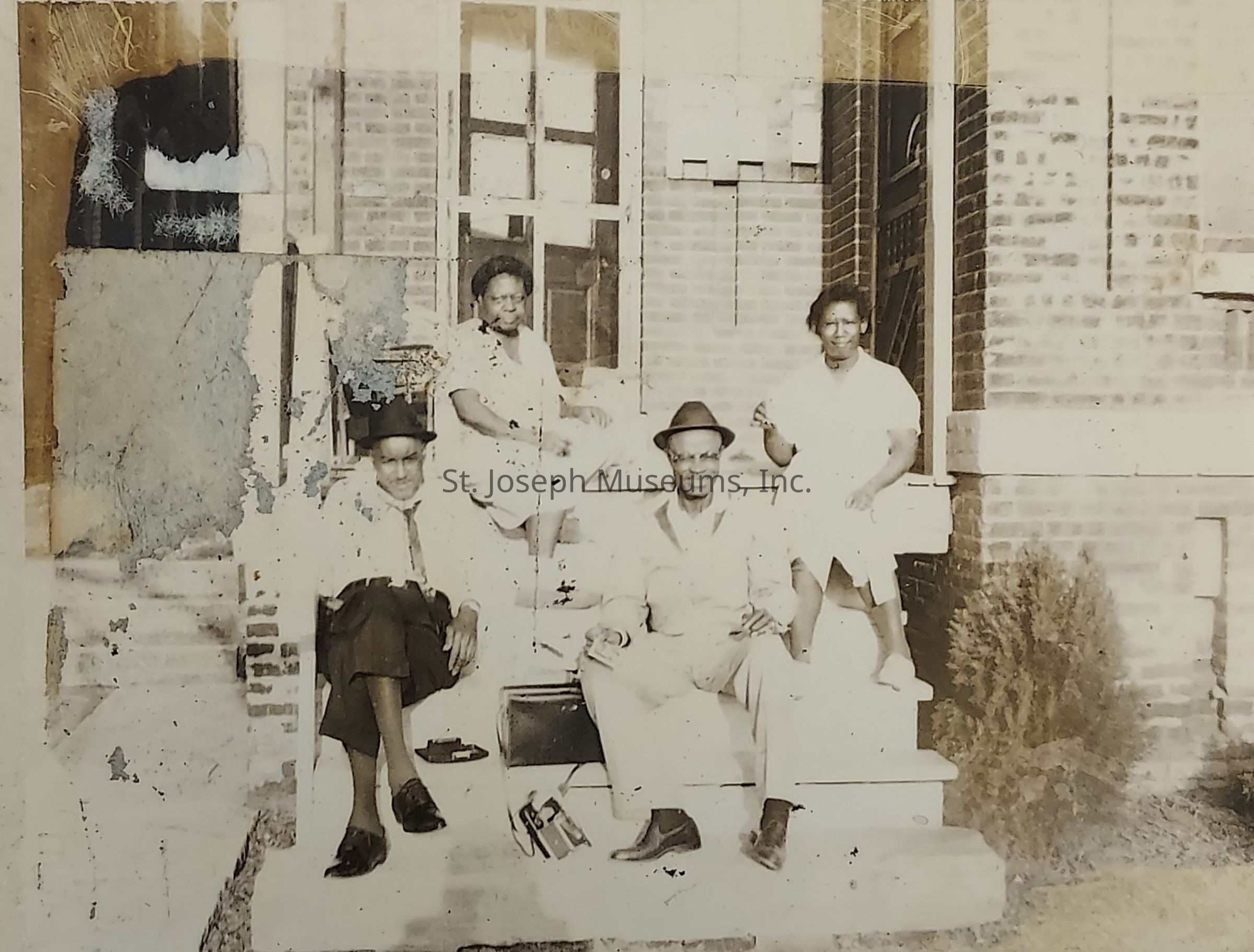 The image depicts four individuals seated on the steps of a brick building, likely taken in the mid-20th century. Two men, sitting on the lower steps, are dressed in suits, with one wearing a hat and the other having a relaxed demeanor. Behind them, two women sit slightly elevated; both are wearing dresses, with one resting her hand on her knee while the other appears comfortable, suggesting a familial or friendly atmosphere. The background shows a detailed brick facade, typical of residential architecture from that era, creating a nostalgic setting. Despite some wear and tear on the photograph, likely due to age, the image captures a moment of camaraderie and tranquility among the group.