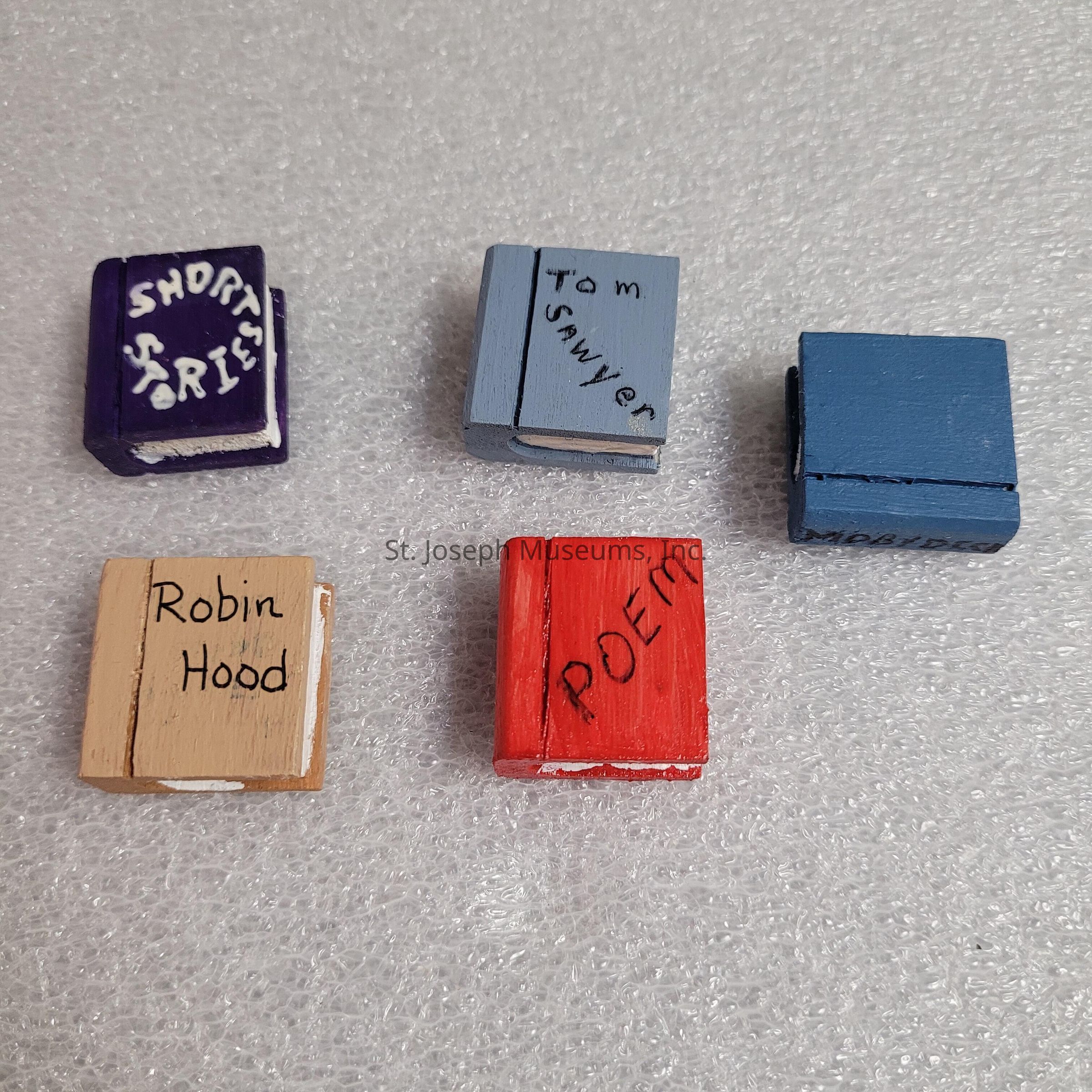 This image shows five small, colorful wooden blocks designed to resemble books, each with a hand-painted title on the cover. The blocks are arranged on a textured white surface. The top left block is purple and labeled "Short Stories," written in a playful white font. Next to it is a light blue block with "Tom Sawyer" written in black script. Adjacent is a solid blue block without visible labeling. In the bottom row, the leftmost block is beige with "Robin Hood" inscribed in black. The last block is bright red, featuring the word "Poem" diagonally across the cover in black letters. These miniature book replicas each appear to convey a classic literary theme, captured in colorful and crafty form, making them both distinctive and decorative objects well-suited for display.