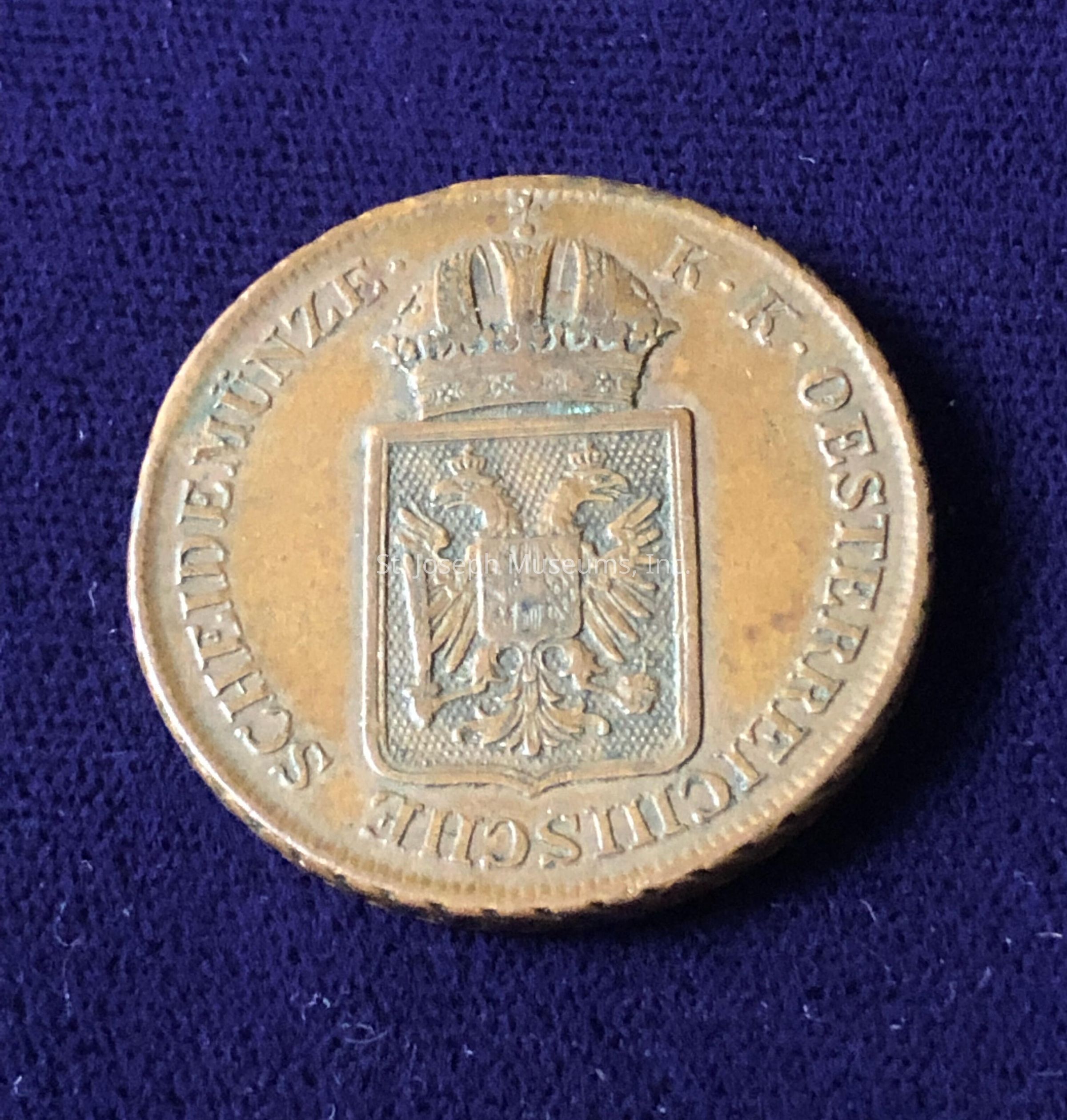 At the center of the coin, there's an emblem with a double-headed eagle, which may represent heraldic significance. Above the emblem is a distinctive crown, suggesting it could be of royal or imperial origin. Along the top edge of the coin, there are inscriptions written in uppercase letters, which read "K. K. OESTERREICHISCHE" and "SCHEIDEMÜNZE," indicating that it pertains to Austrian currency, possibly from the Austro-Hungarian period. The coin's surface exhibits signs of age, with patches of tarnish and patina.