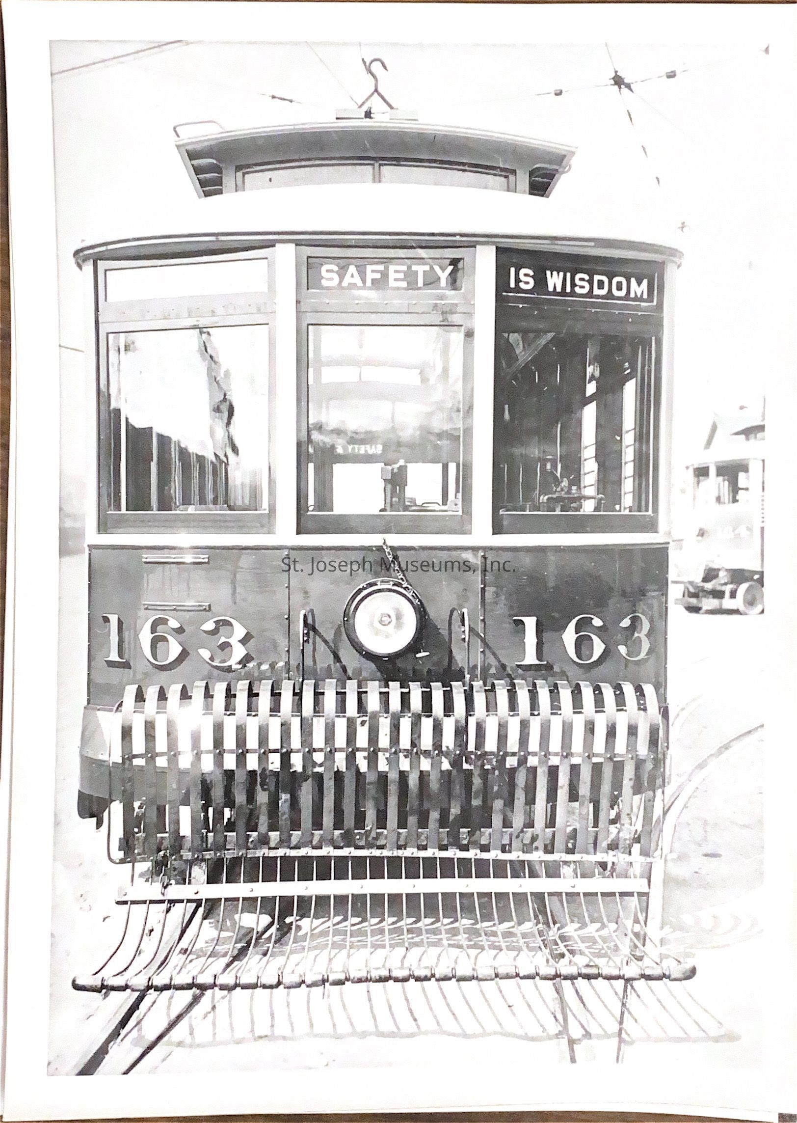 Black and white photograph featuring the front of an old tram car with the number 163 prominently displayed on both sides of a central headlight, and above the windows the phrase "SAFETY IS WISDOM" is written, while below, a metal guard is attached to the tram, possibly serving as a protective barrier; the background reveals some architectural structures and parts of another tram.