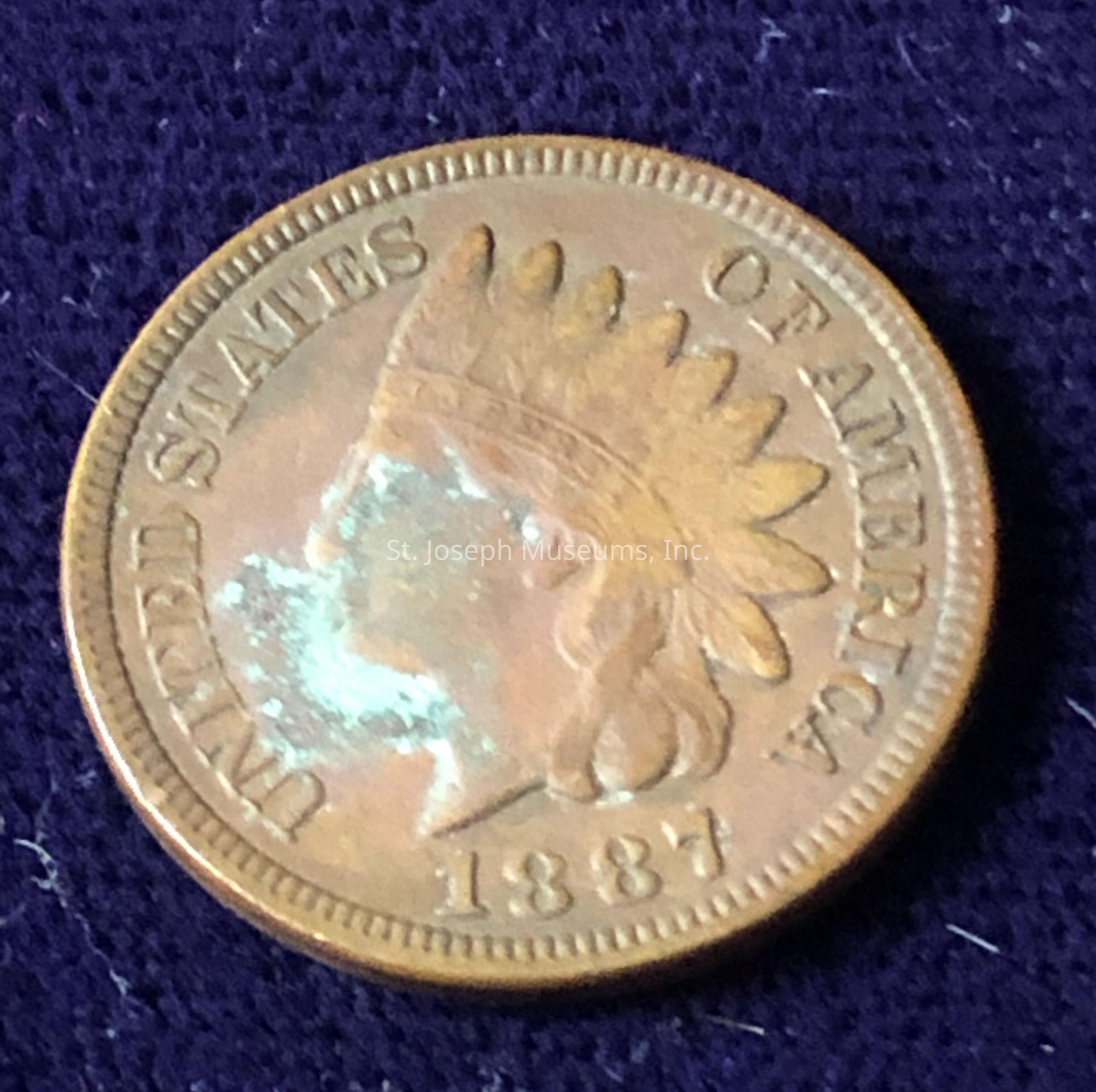 The image shows an 1887 Indian Head penny from the United States, featuring the profile of Lady Liberty wearing a Native American headdress. The text "UNITED STATES OF AMERICA" and the date are inscribed around the edge. The coin, made of bronze, rests on a dark textured fabric, and while it shows signs of age, including some tarnishing and wear, the details of the design remain relatively clear, highlighting the craftsmanship of the era. Significant for collectors, this coin represents a piece of American history, often admired for its artistic depiction and historical significance, reflecting the nation's past economic practices.