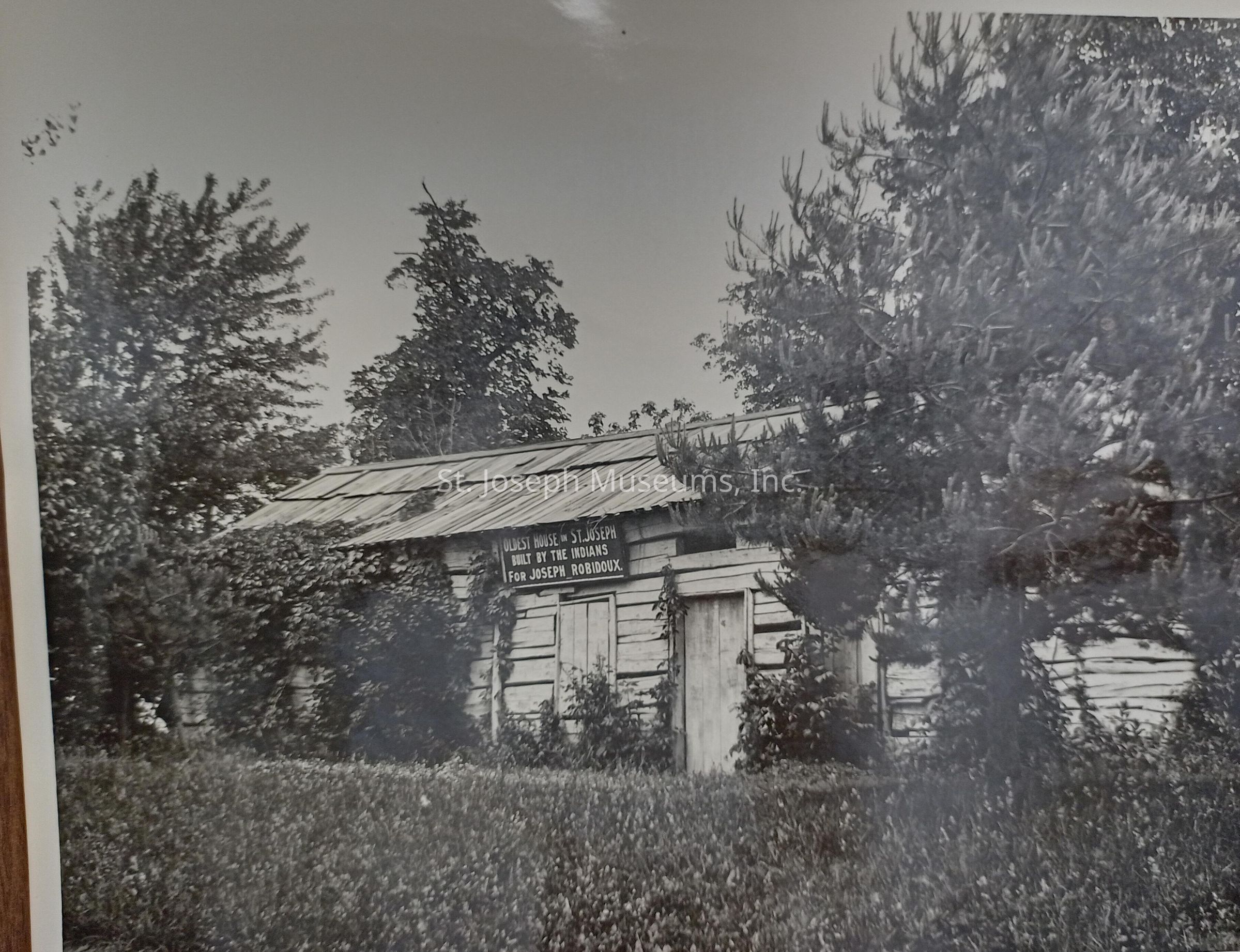A black-and-white photograph depicting an old wooden structure claiming to be the "oldest house in St. Joseph," built by a Native American tribe for Joseph Robidoux, according to the sign on the building. Surrounded by dense vegetation and trees, the rustic log cabin stands with a metal roof, blending into the natural environment. The house, partially obscured by foliage, appears weathered.