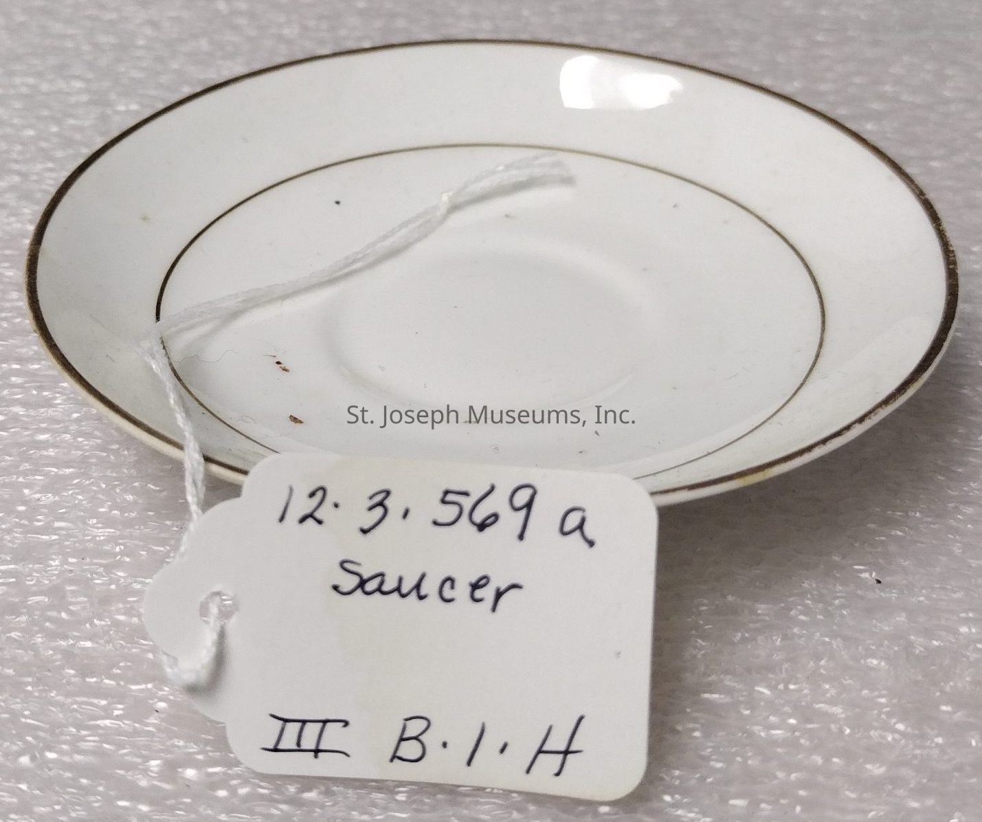 The image shows a white porcelain saucer with a delicate, thin gold rim around its edge, resting on a textured, light-colored surface. In the center of the saucer is a slight depression, typical of its design to hold a teacup. Attached to the saucer is a white string with a label that appears to be made of card or paper. The label has handwritten text in dark ink, which includes the date or code “12.3.569a” along with the word “Saucer” and additional characters or markings, “III B.1.H.” The saucer is simple yet elegant, suggesting it might be a piece from a larger set, possibly vintage or antique. The lighting in the image is soft, casting a mild reflection on the saucer’s glossy surface, highlighting its smooth, pristine condition, free of any visible blemishes or chips, aside from slight discoloration or marks near the center. This saucer is likely cataloged for documentation, collection,
