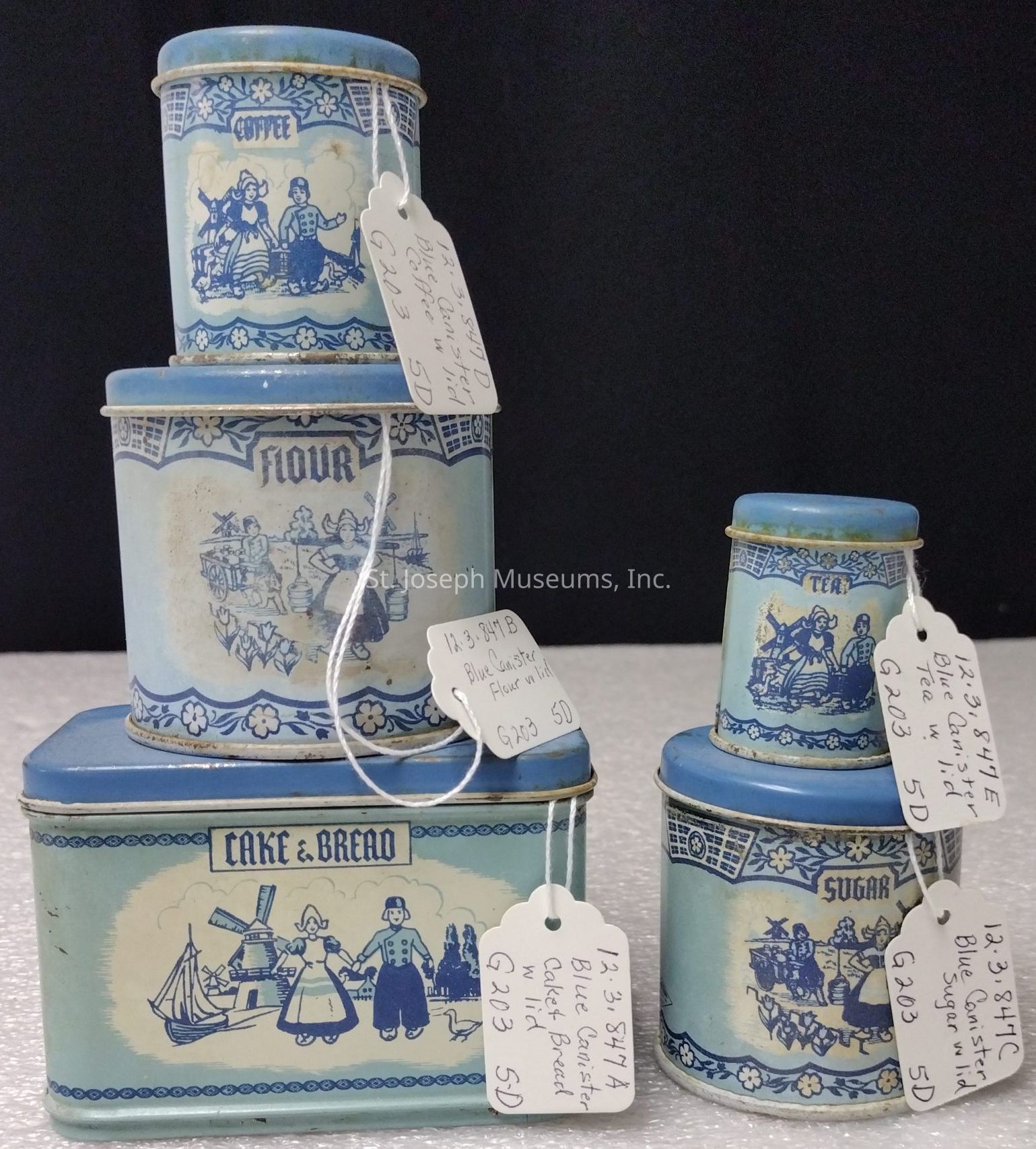 The image displays a set of vintage blue canisters, each adorned with intricate and nostalgic blue-and-white designs reminiscent of Dutch or traditional European motifs. These canisters, labeled for storing different kitchen staples such as coffee, flour, tea, cake & bread, and sugar, are stacked next to each other, showcasing their different sizes. Each canister is adorned with scenes featuring figures in traditional attire and idyllic settings like windmills and boats. Attached to each canister is a price tag with handwritten notes, hinting at their collectible status or sale in an antique setting. The aesthetic is quaint and charming, evoking a sense of rustic elegance and a time gone by.