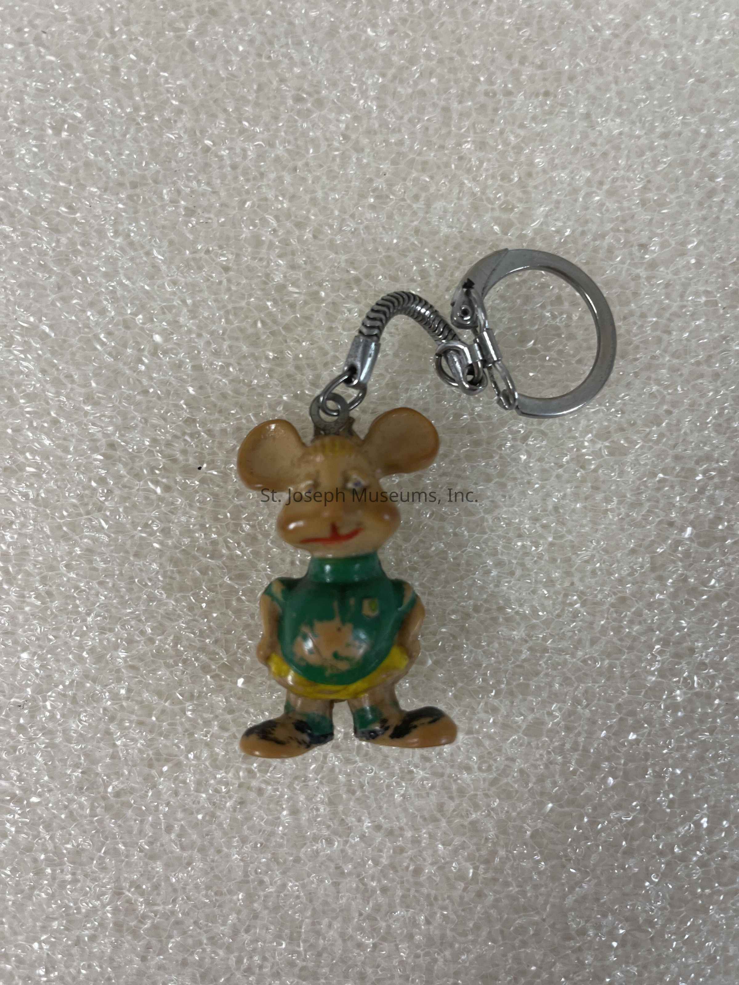The image shows a small keychain featuring a cartoonish mouse figure with round ears and a smiling expression. The mouse is wearing a green shirt and yellow shorts, and it stands upright with its arms relaxed by its sides. The keychain attachment is metallic, with a small chain connecting the mouse to a circular keyring for easy attachment to keys or bags. The background consists of a textured white material that closely resembles bubble wrap or foam, creating a soft contrast with the glossy and colorful finish of the mouse figure. The combination of the character's playful appearance and the functional keyring design makes this accessory both charming and practical for everyday use, appealing to those who appreciate whimsical and cute items.