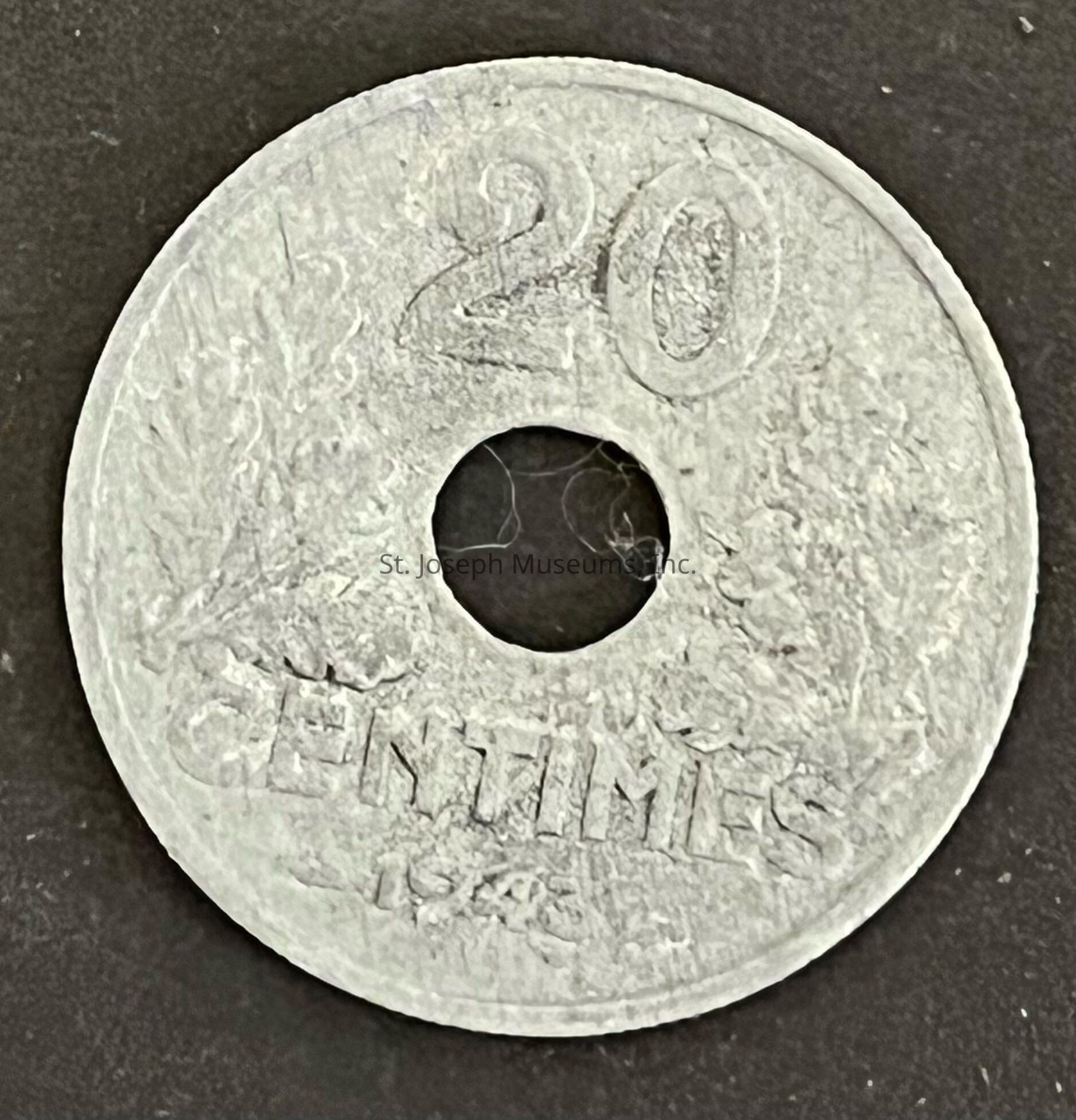 The image displays a vintage 20 centimes coin with a distinctive circular hole in the center, set against a dark background. The coin is made of a metal that appears somewhat tarnished or worn, suggesting age and historical usage. The denomination "20" is clearly inscribed at the top, with "CENTIMES" prominently labeled beneath it. The year "1945" is engraved at the bottom, marking its minting period as the mid-20th century. The coin features intricate decorative elements, including faint foliage patterns along the sides, providing insight into its design aesthetics. The overall appearance of the coin, with its unique central hole and historical inscription, hints at its potential origin as a piece of currency used in France or a French-speaking region during the post-World War II era. The wear and patina visible on the coin's surface suggest it has been circulated extensively, carrying with it a sense of history and the passage of time.