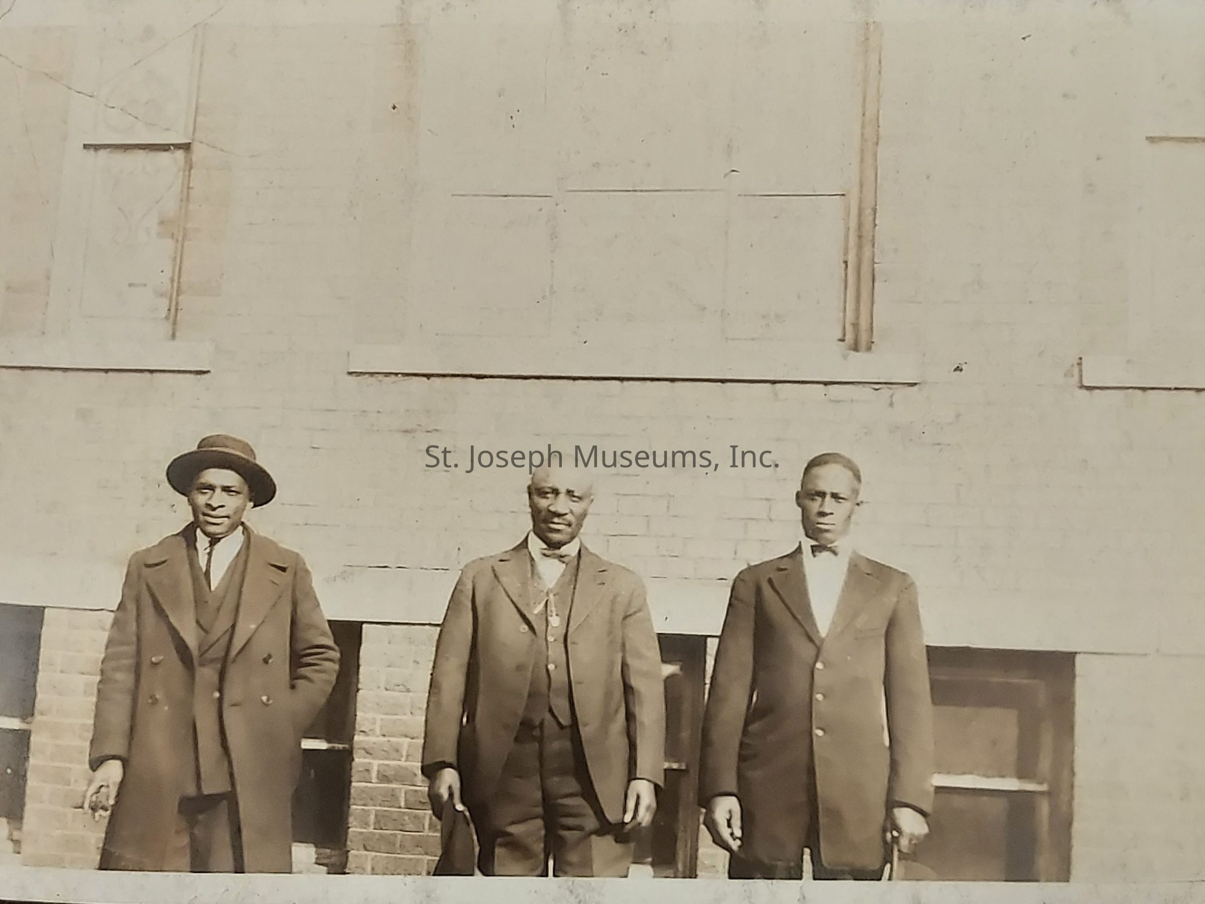 The image depicts three men standing side by side in front of a brick building, each wearing formal suits that likely represent the fashion of the early 20th century, evident from their long overcoats and dress shoes. The man on the left is wearing a hat and a double-breasted coat, while the middle man, who appears older, has thinning hair and is holding a book or document in one hand, his slightly more relaxed posture suggesting an approachable demeanor. The man on the right is dressed in a single-breasted suit and a bow tie, maintaining a formal and upright stance, with all three facing the camera directly, their expressions conveying a sense of composure and dignity. The building behind them features closed windows, providing a backdrop that complements the overall solemn and respectful tone of the photograph, which evokes a sense of historical significance and cultural heritage.