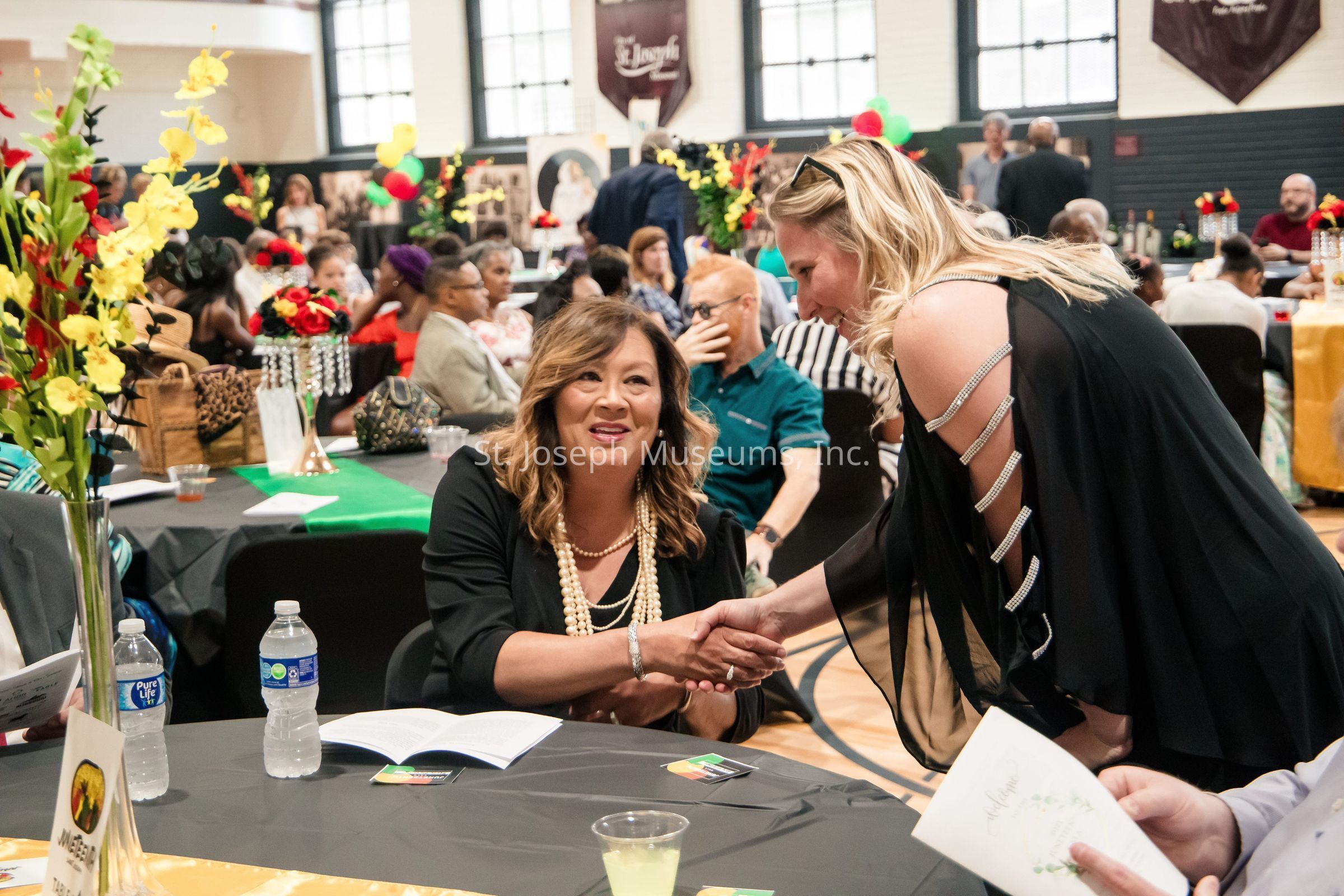 In a lively and colorful indoor setting, two women engage warmly, one seated at a table with a notebook and water bottle, and the other standing, leaning in to shake hands while holding a program. The room is bustling with people seated at tables covered in green, black, and gold tablecloths, adorned with vibrant floral arrangements, creating a festive atmosphere. Behind them, a variety of decor, including balloons and framed portraits, adds to the celebratory ambiance. The interior features large windows and brown banners on the walls, suggesting a venue like a community hall or banquet space.