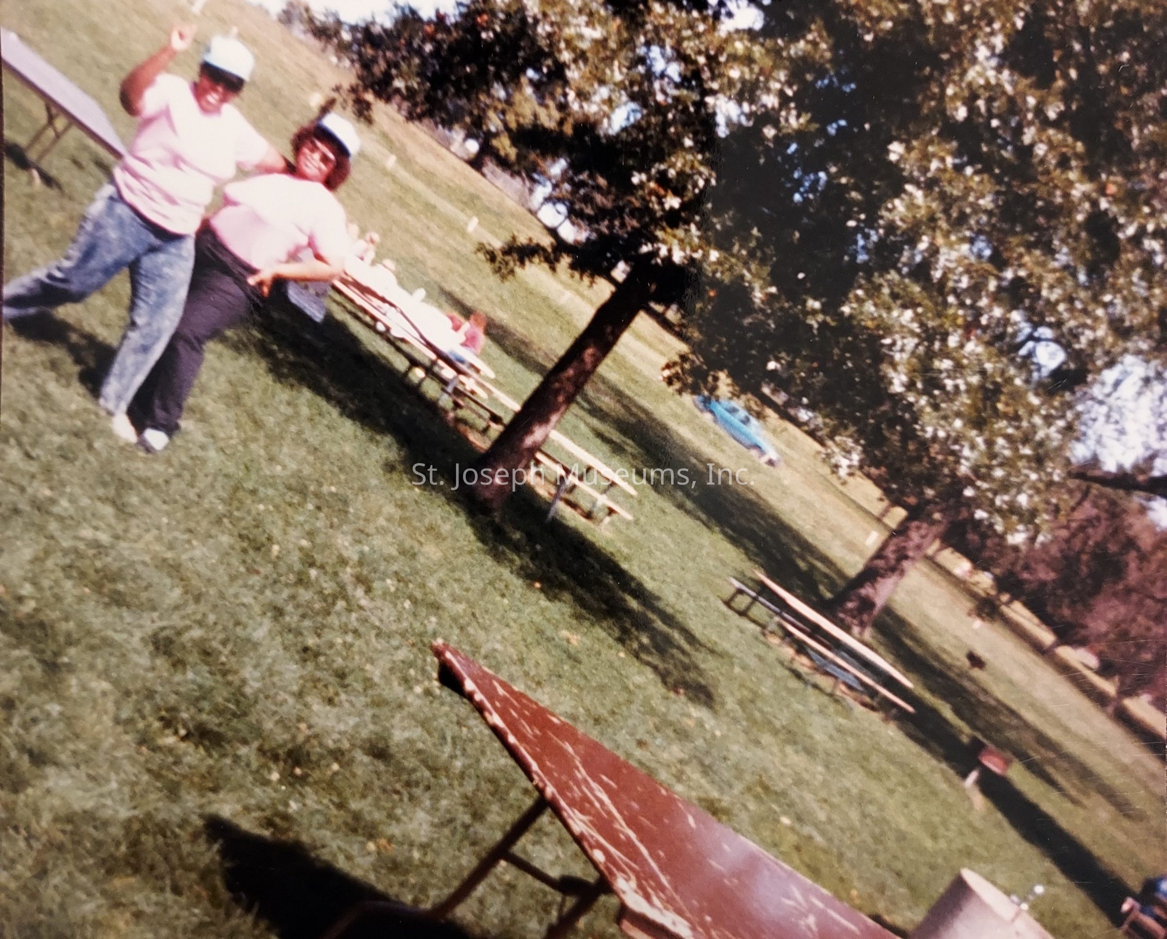 In the image, two people are joyfully posing on a grassy field, wearing matching light-colored shirts and baseball caps; they are standing near a wooden picnic table under the shade of a large tree. The setting suggests a relaxed outdoor gathering or picnic at a park, visible through scattered picnic tables and people in the background enjoying the sun. The atmosphere appears warm and sunny, with shadows cast on the vibrant green grass, while the distant horizon shows more trees under a clear sky, creating an inviting and cheerful scene.